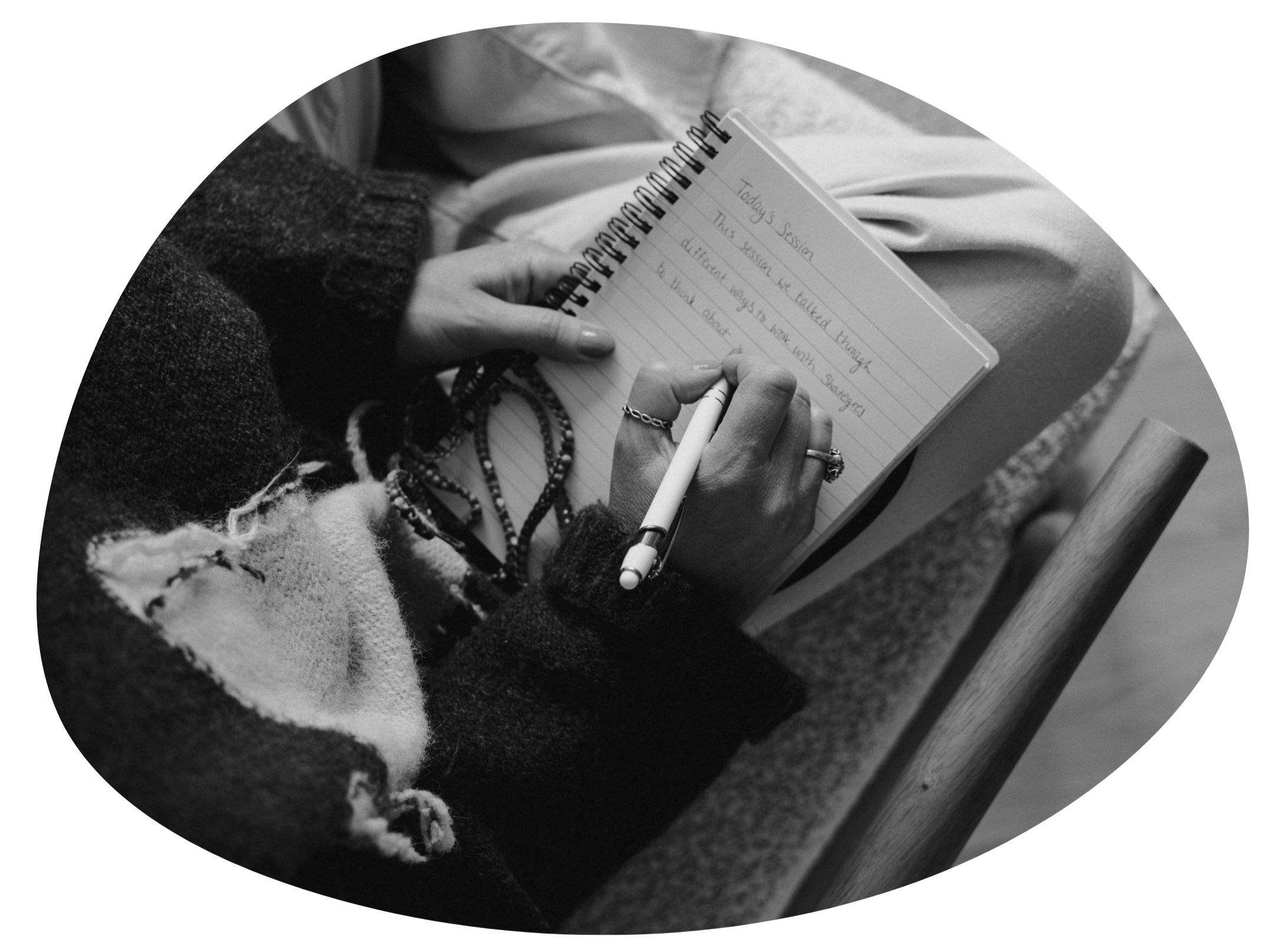 A person holding a spiral notebook while writing with a pen, sitting on a chair.