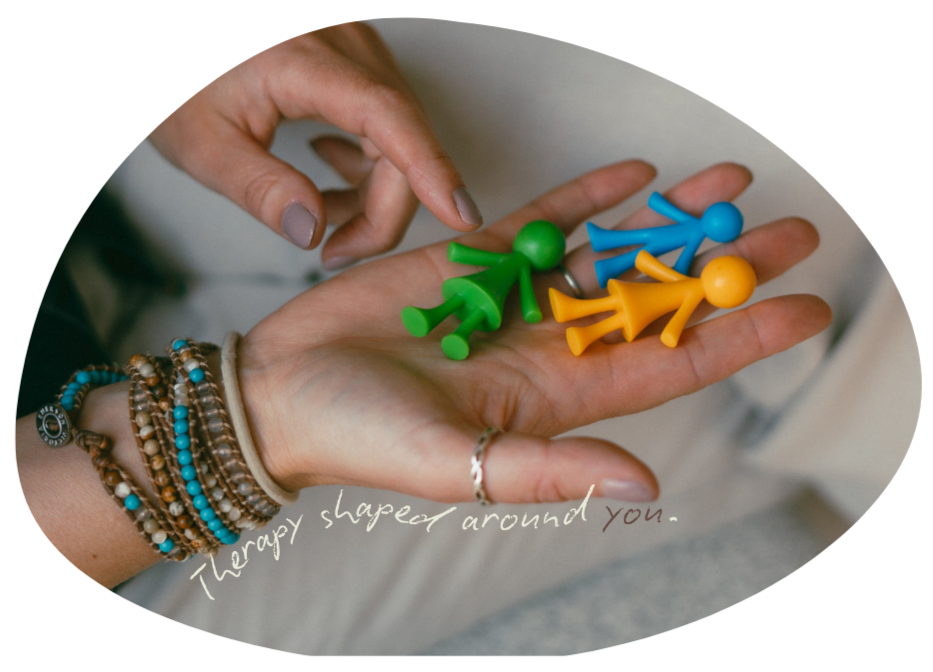 A person's hand holding colorful plastic figurines in green, blue, and yellow, with handwritten text reading "Therapy shaped around you."