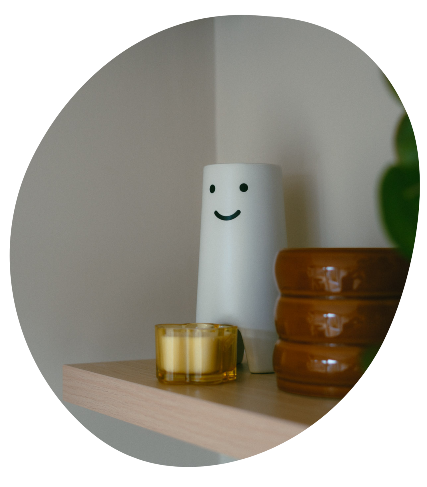 Decorative shelf with a white vase featuring a smiling face, a yellow candle, a brown ceramic bowl, and a small green plant.