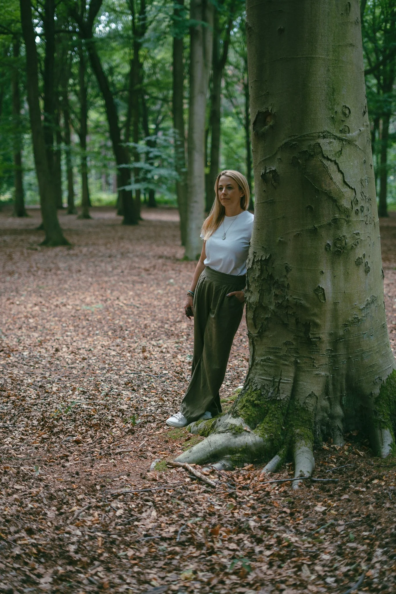 A woman with long blonde hair wearing a white t-shirt, green pants, and white sneakers, standing in a forest, leaning against a large tree with her hands in her pockets.