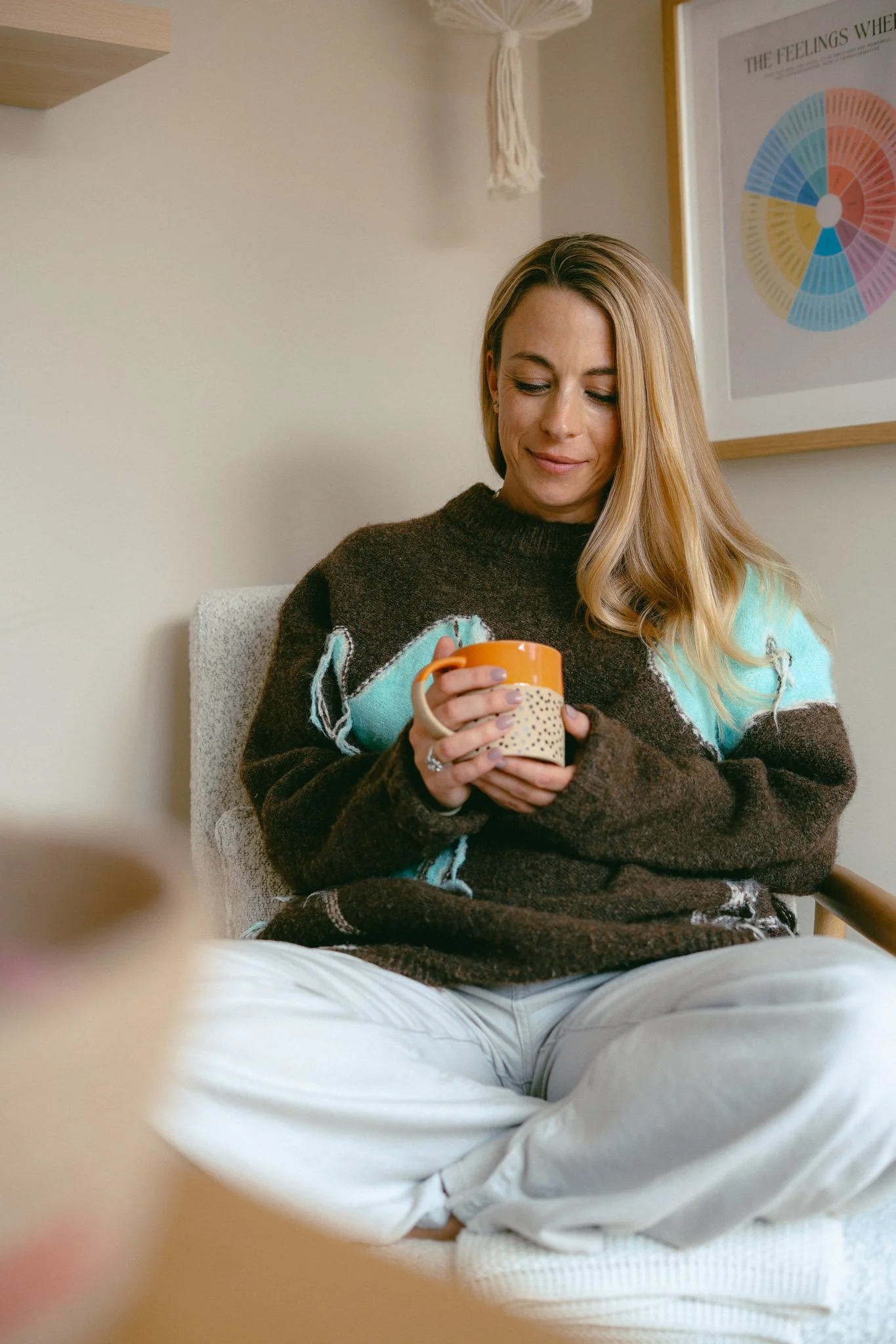 A woman with blonde hair sitting in a chair, holding an orange mug, wearing a brown sweater, and smiling softly. There is a color wheel poster on the wall behind her.