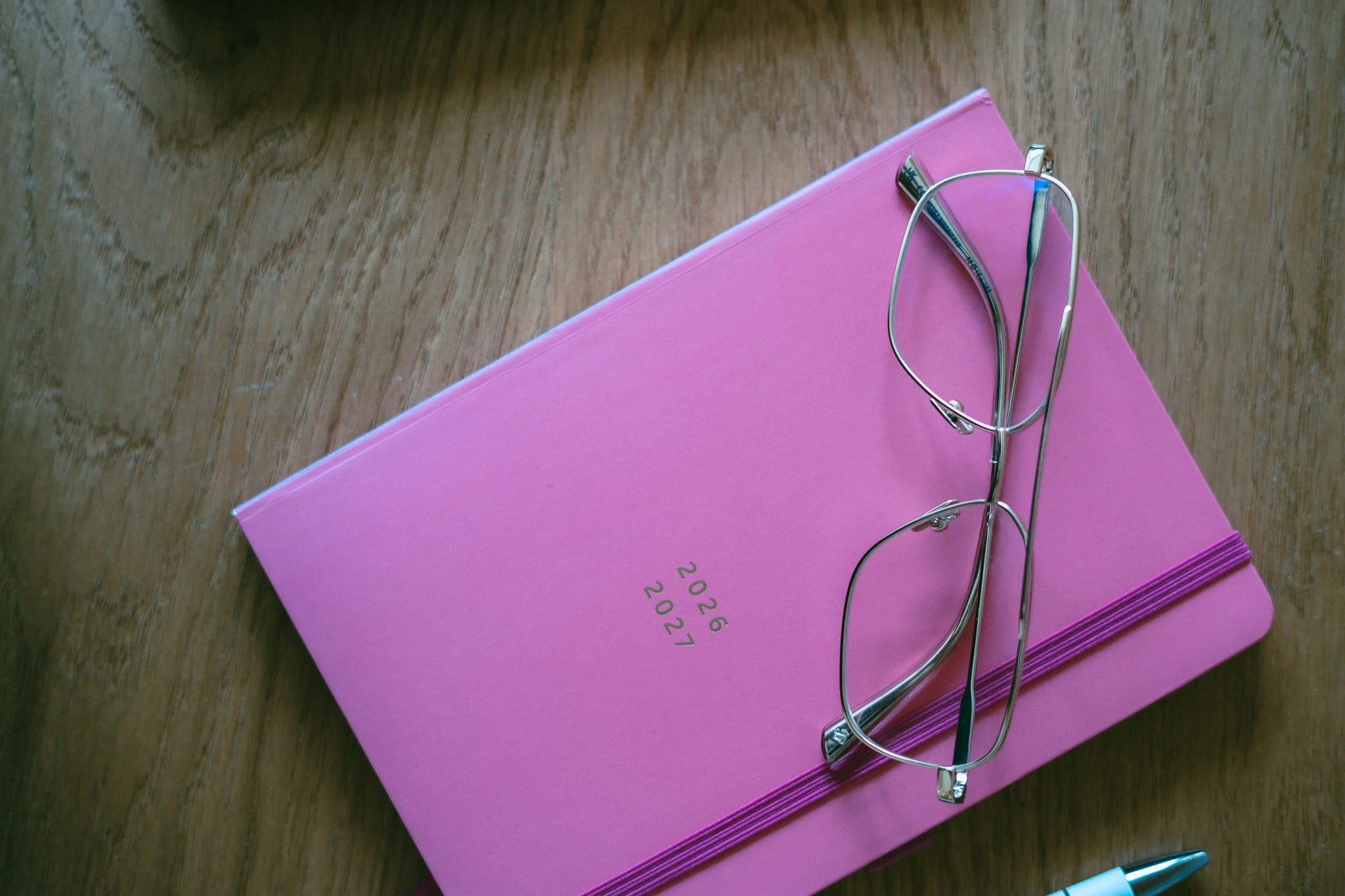 A pink 2024 planner with gold text, a pair of glasses, and a white pen on a wooden surface.