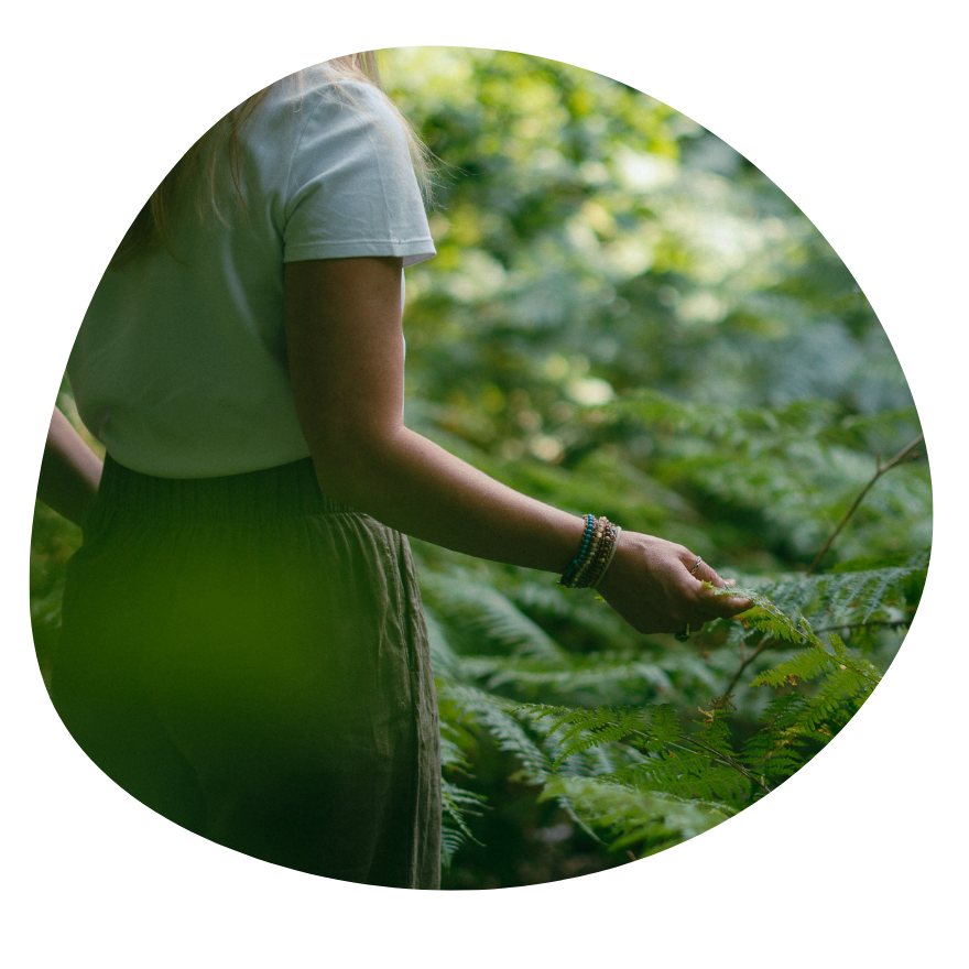 A person with long hair wearing a white T-shirt and a skirt standing in a lush green outdoor setting, gently touching plants.