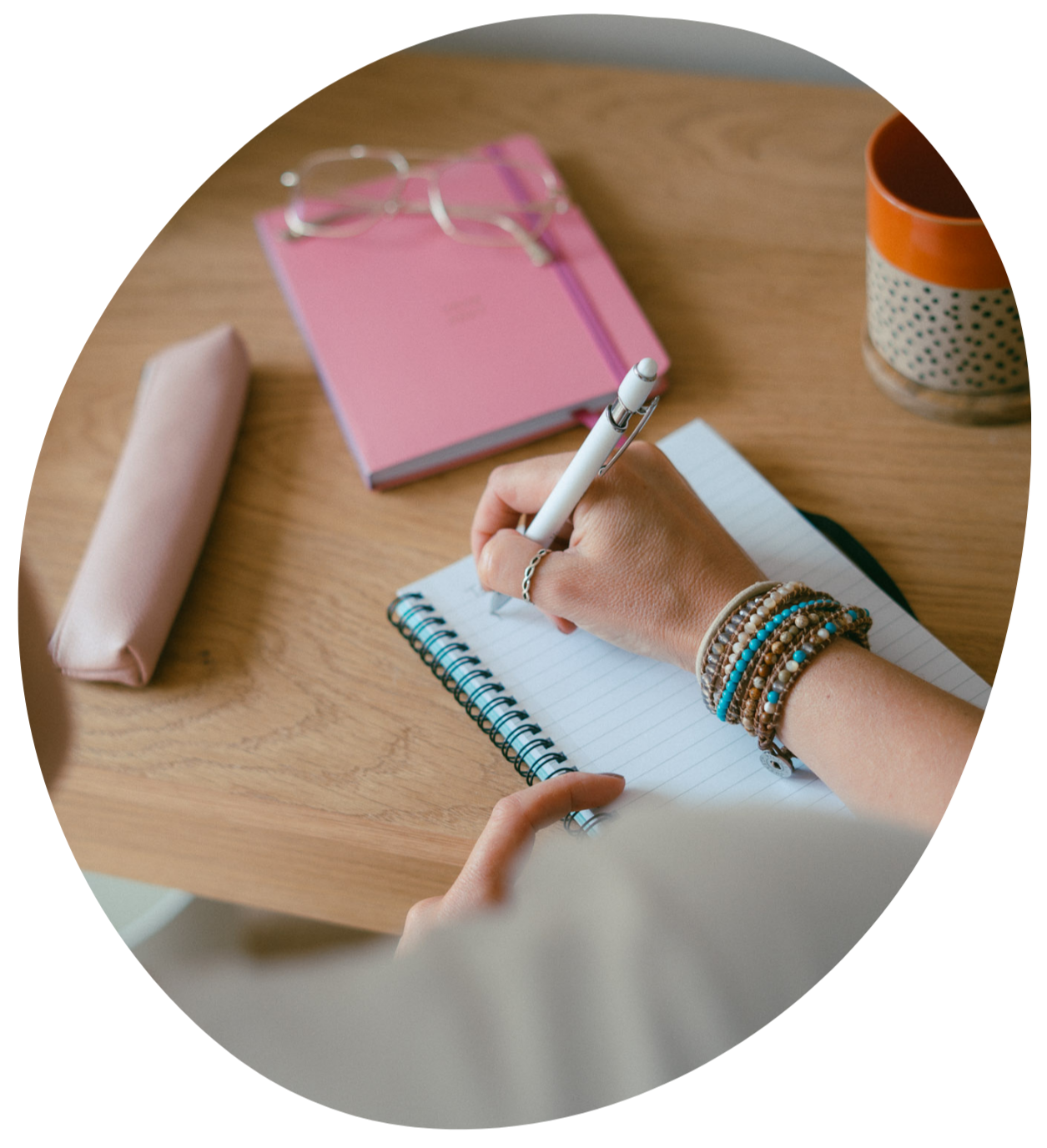 Person writing in a spiral notebook on a wooden desk, with a pink notebook, glasses, a pencil, a pink pencil case, a ceramic cup, and bracelets on their wrist.