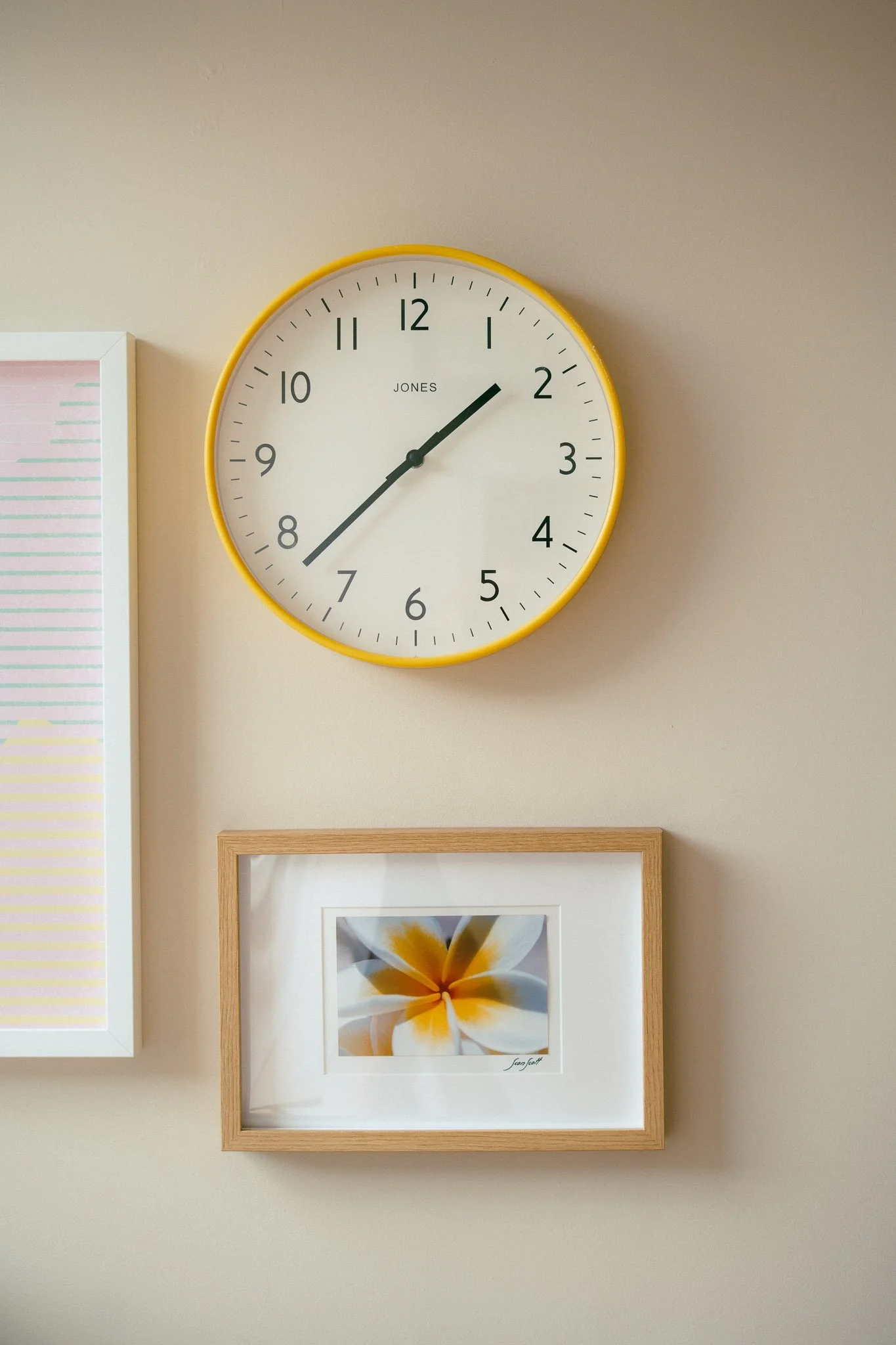 A wall clock with a yellow rim shows the time as 1:11, mounted above framed artwork of a flower with white and yellow petals.
