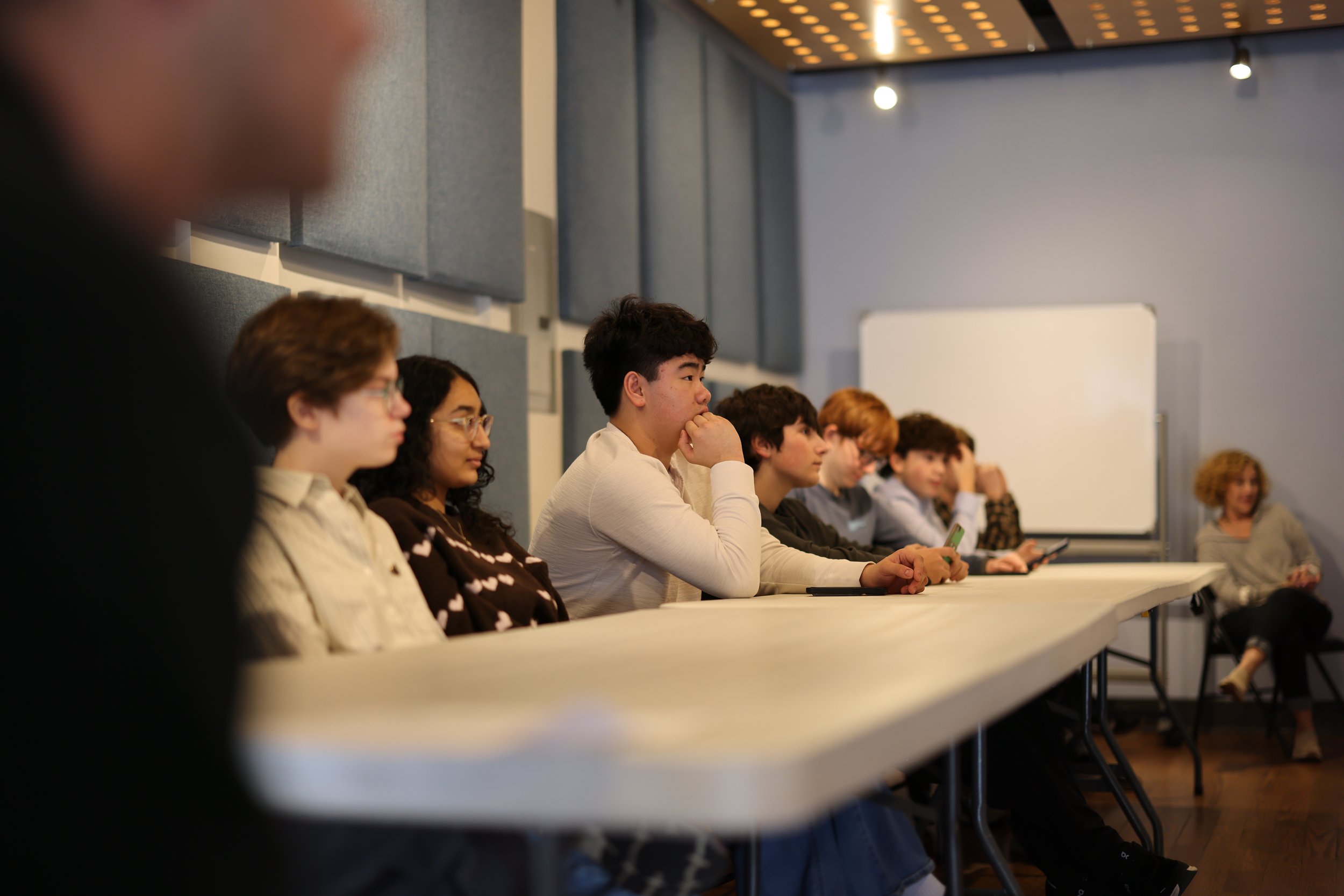A diverse group of people seated at a long table, attentively listening during a presentation or lecture in a modern conference room with gray acoustic panels on the walls and a whiteboard in the background.
