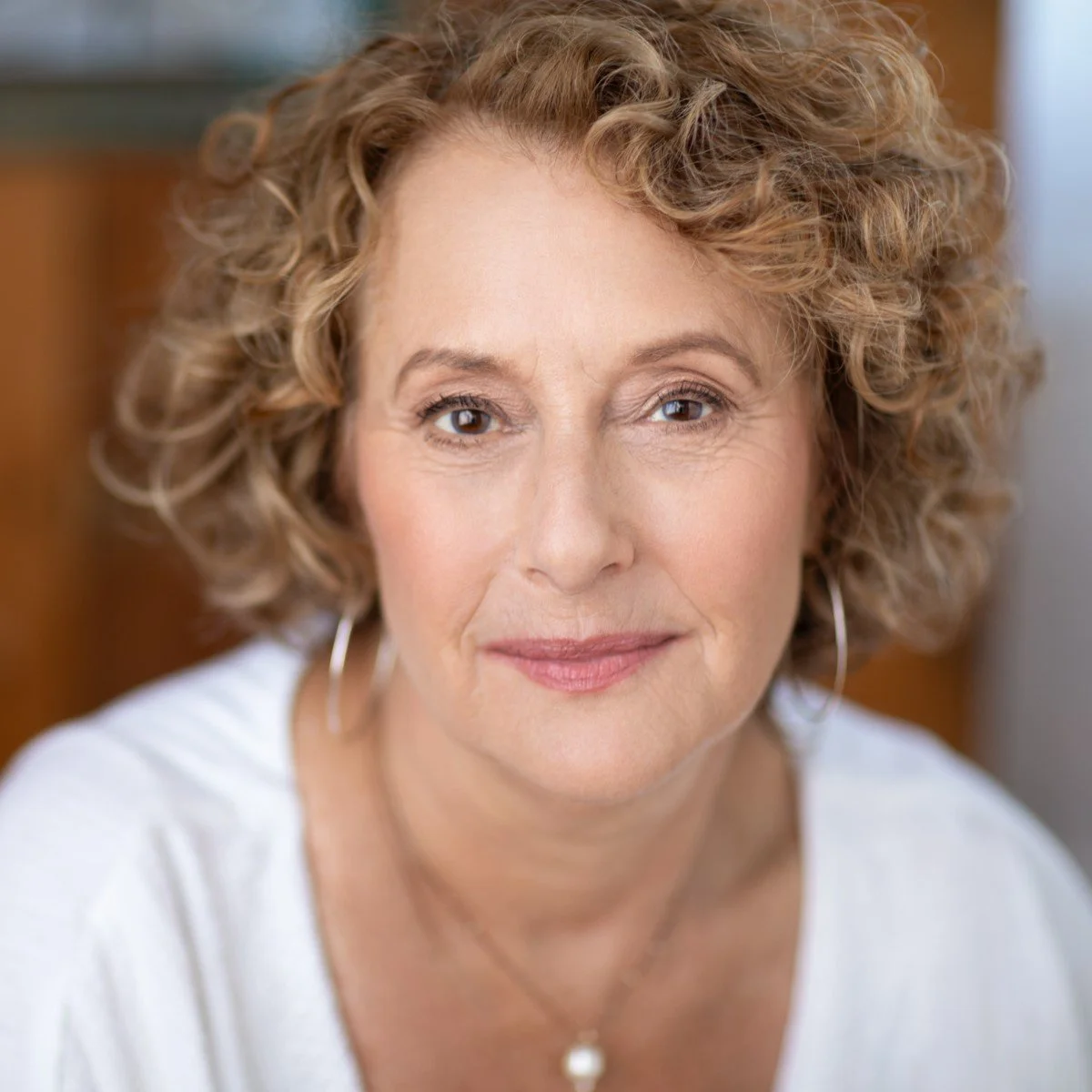 Close-up of a middle-aged woman with curly blonde hair, wearing earrings and a white top, smiling softly at the camera.