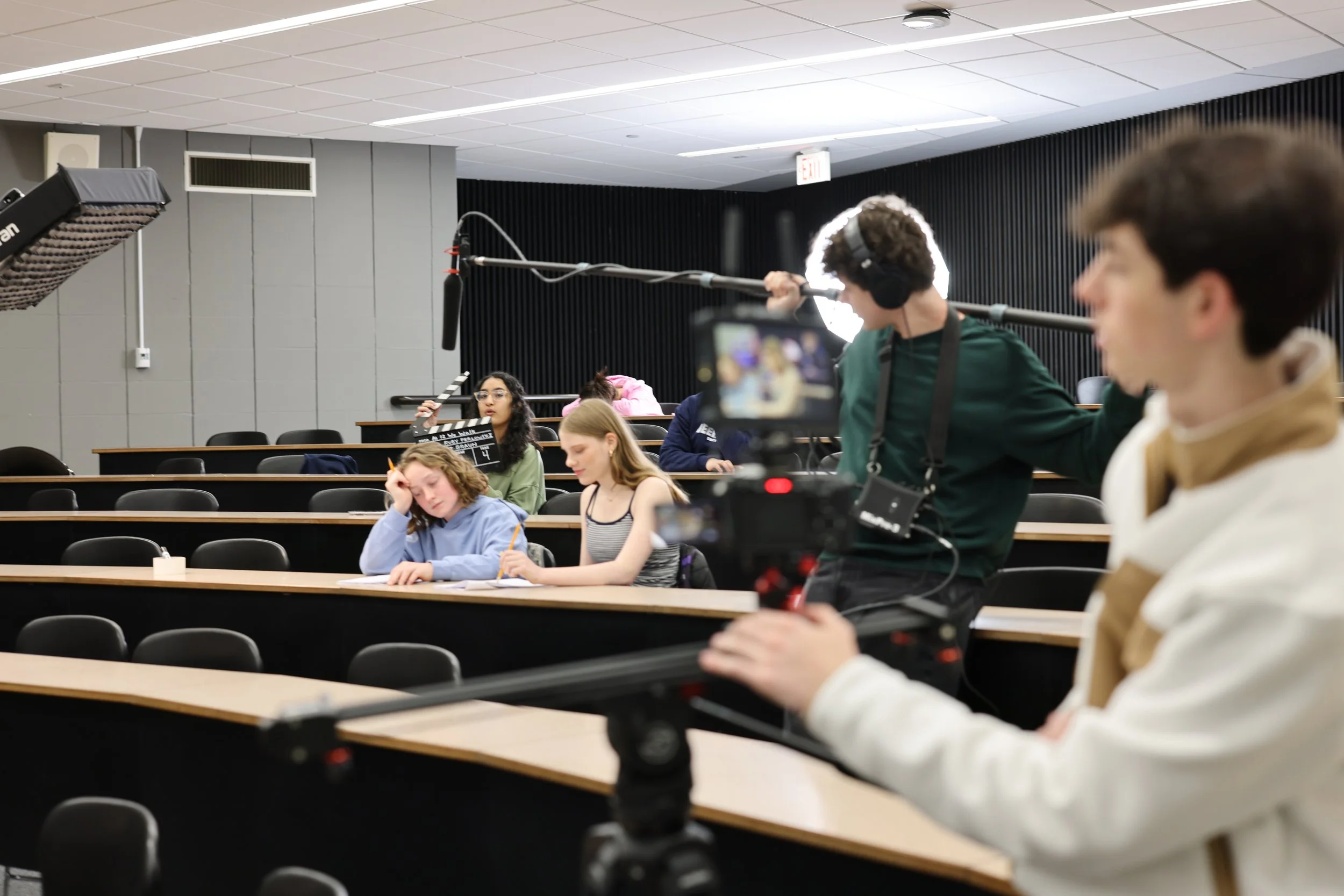 A classroom scene with students seated at desks, some taking notes, and a person in the foreground filming with a professional camera, while another person operates a boom microphone overhead.