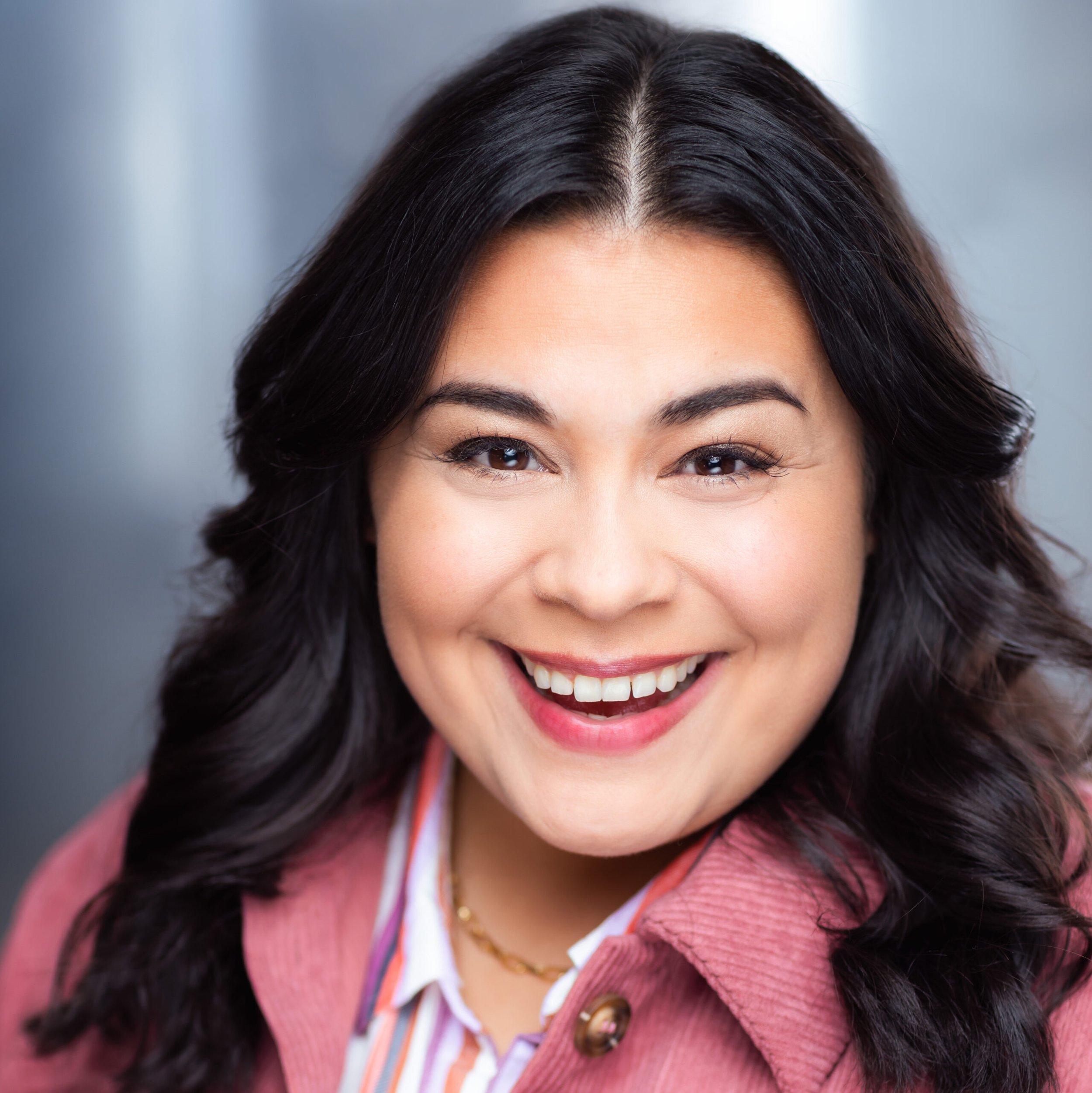 Close-up of a woman's face smiling, showing her eyes, nose, and mouth, with black wavy hair and wearing a pink jacket over a white shirt with orange, purple, and pink stripes.