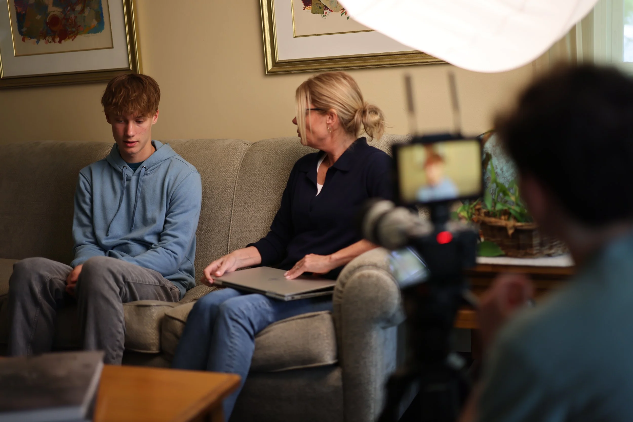 A young man in a light blue hoodie sitting on a beige sofa with a woman with glasses, blonde hair tied in a bun, and a dark jacket, engaging in conversation. A person in the foreground is filming or taking a photo of them with a camera, and large framed artwork hangs on the wall behind.