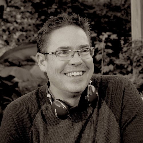 A smiling man with glasses and headphones around his neck, outdoors with foliage in the background.