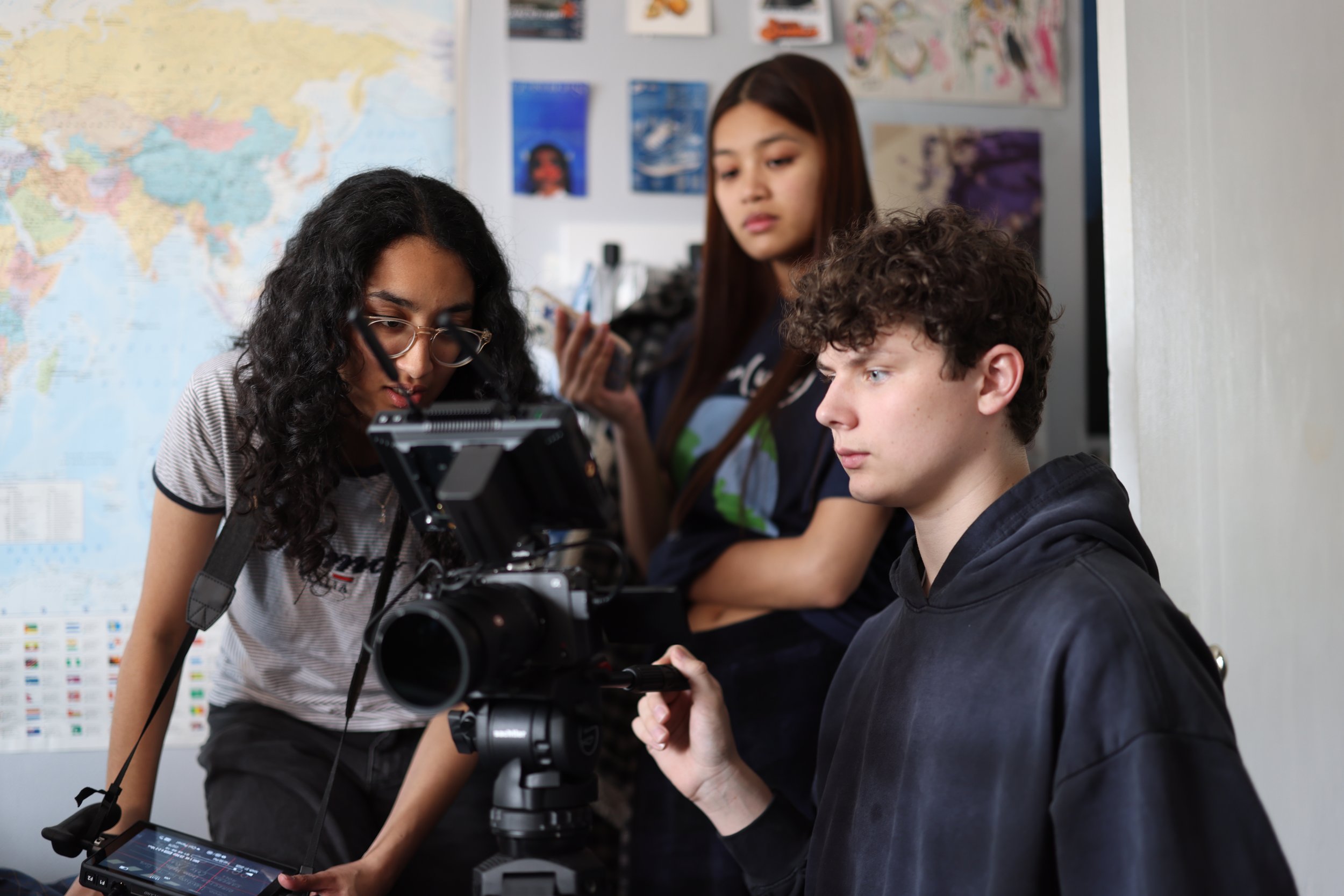 Three students in a classroom examine a camera mounted on a tripod, with a large world map on the wall behind them and various posters and artwork.
