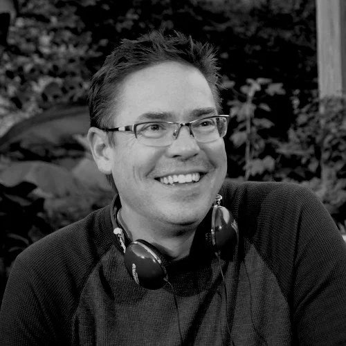 A smiling man wearing glasses and headphones around his neck, outdoors with trees in the background.