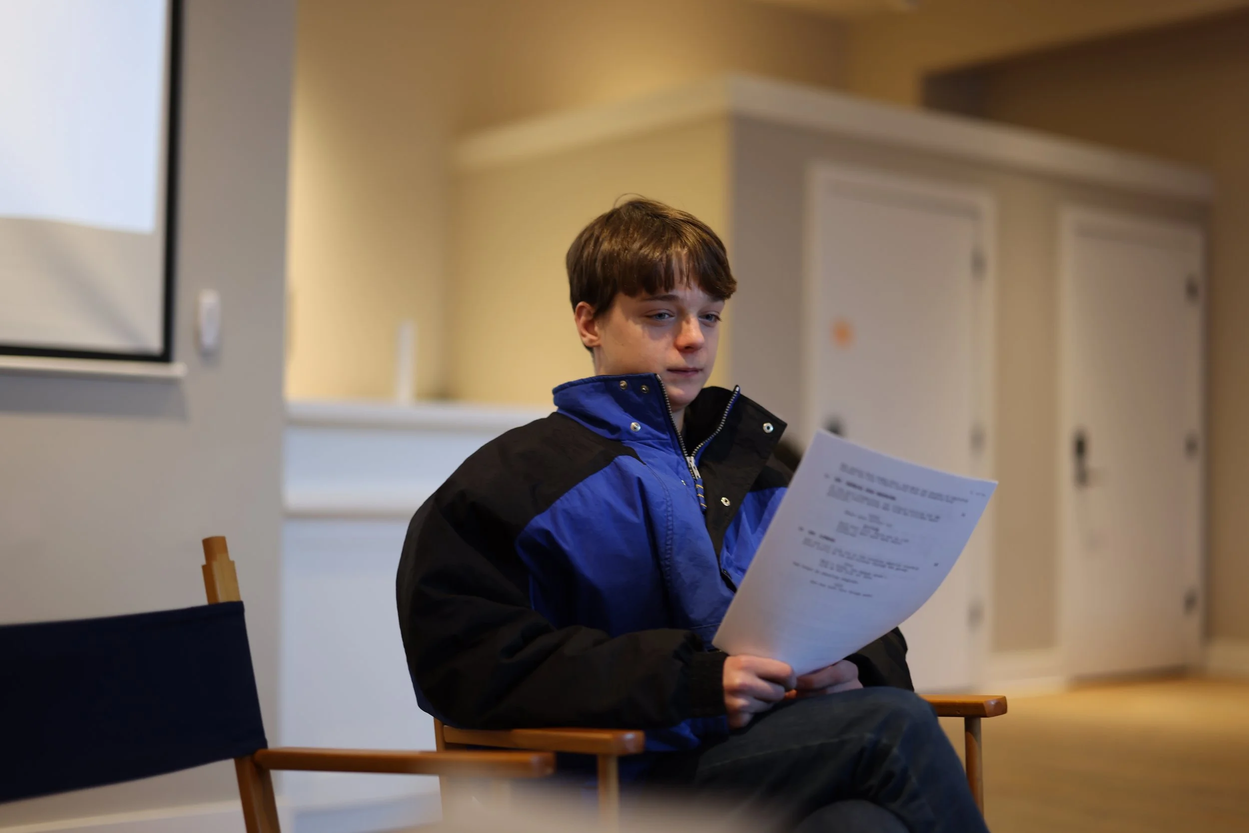 A young boy with brown hair sitting on a wooden chair indoors, wearing a blue and black jacket, holding and looking at a sheet of paper