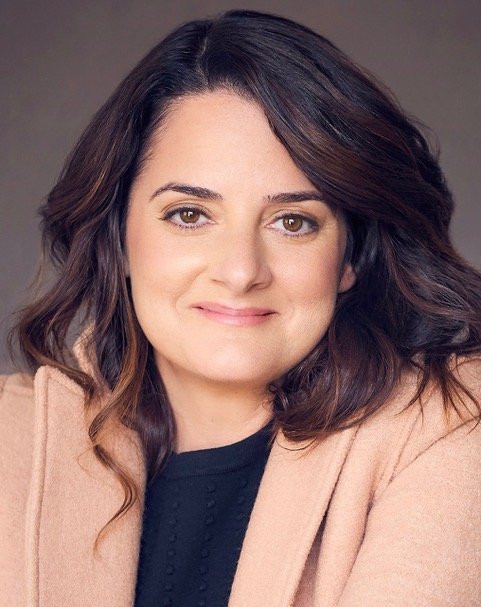 Close-up of a woman with wavy dark brown hair, wearing a beige blazer and a black top, smiling softly at the camera.