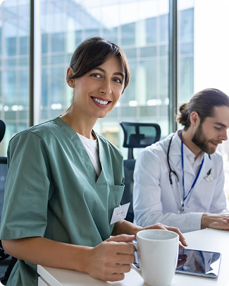 A female healthcare worker smiling and holding a coffee mug in a clinical setting with a male nurse working at a desk in the background.