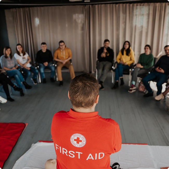 A person in a red shirt with a Red Cross logo and the words 'First Aid' on the back is teaching a group of young adults in a circle in a room with gray curtains. The group is seated around them, listening attentively.
