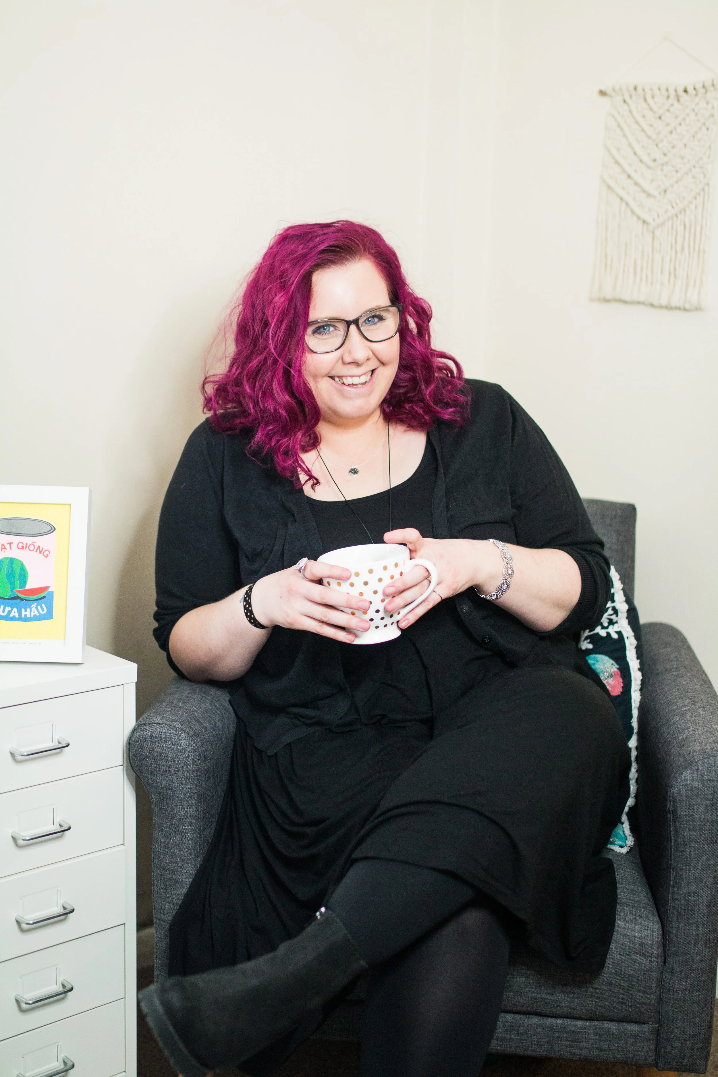 Cariad Martin, a woman with pink hair, glasses, and a black dress sitting in a gray armchair, holding a white mug with gold polka dots and smiling at the camera.