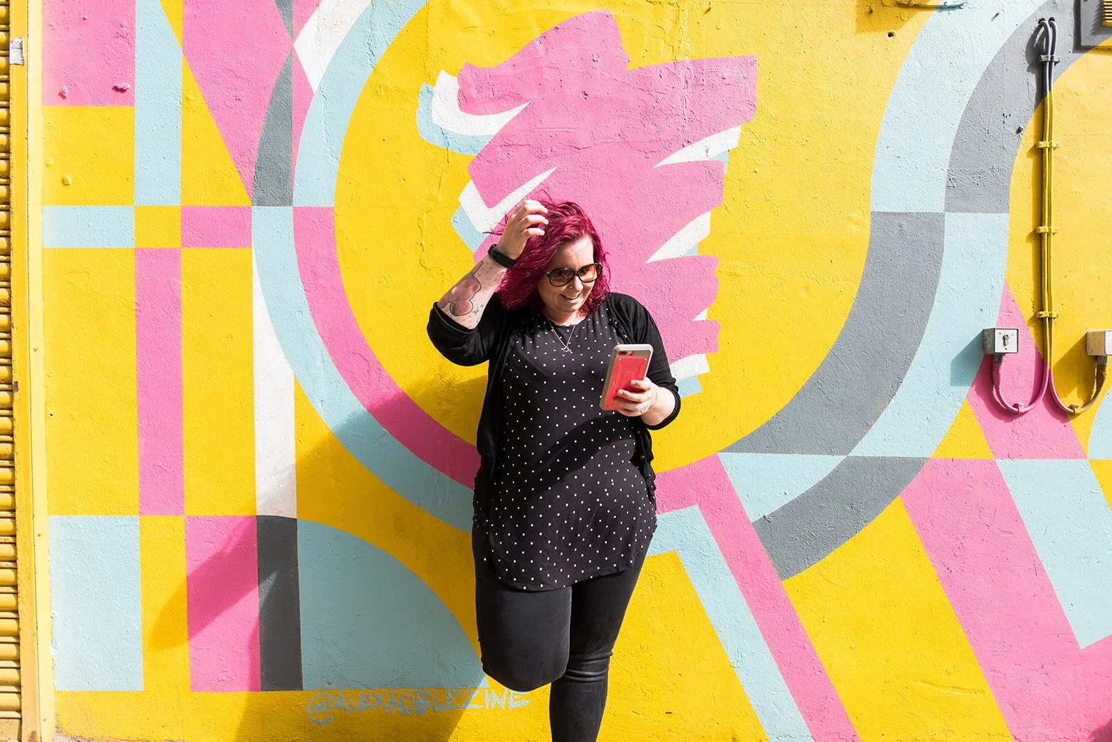 Cariad Martin, a woman with pink hair, glasses, and tattoos is standing in front of a colorful yellow and pink graffiti mural while looking at her phone.