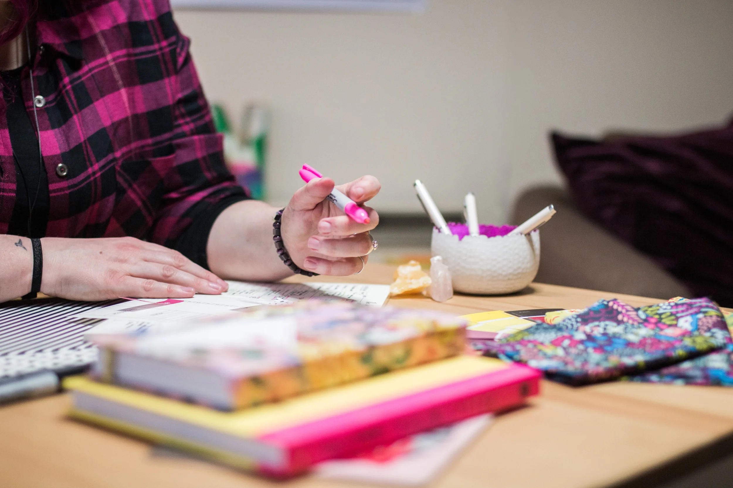 Person in a red and black plaid shirt writing with a pink marker at a cluttered table with various papers, art supplies, and a white bowl filled with pink items.