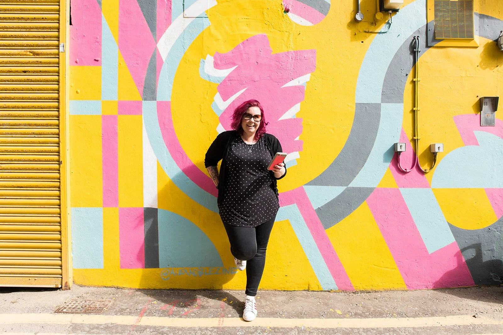 Cariad Martin, a woman with pink hair and sunglasses smiling while standing in front of a colorful abstract mural on a yellow wall, holding a smartphone.