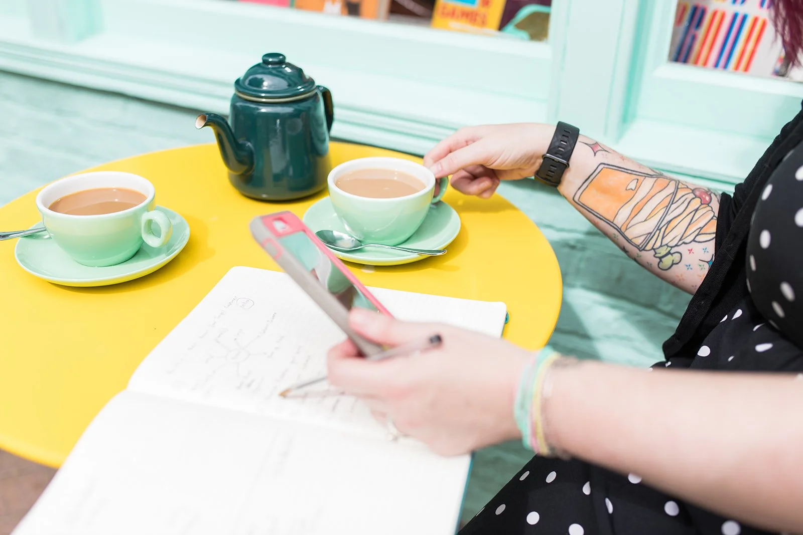 A person with a tattooed arm and polka dot dress sitting at a yellow table with two cups of tea, a small teapot, a spoon, a notebook, and a smartphone.