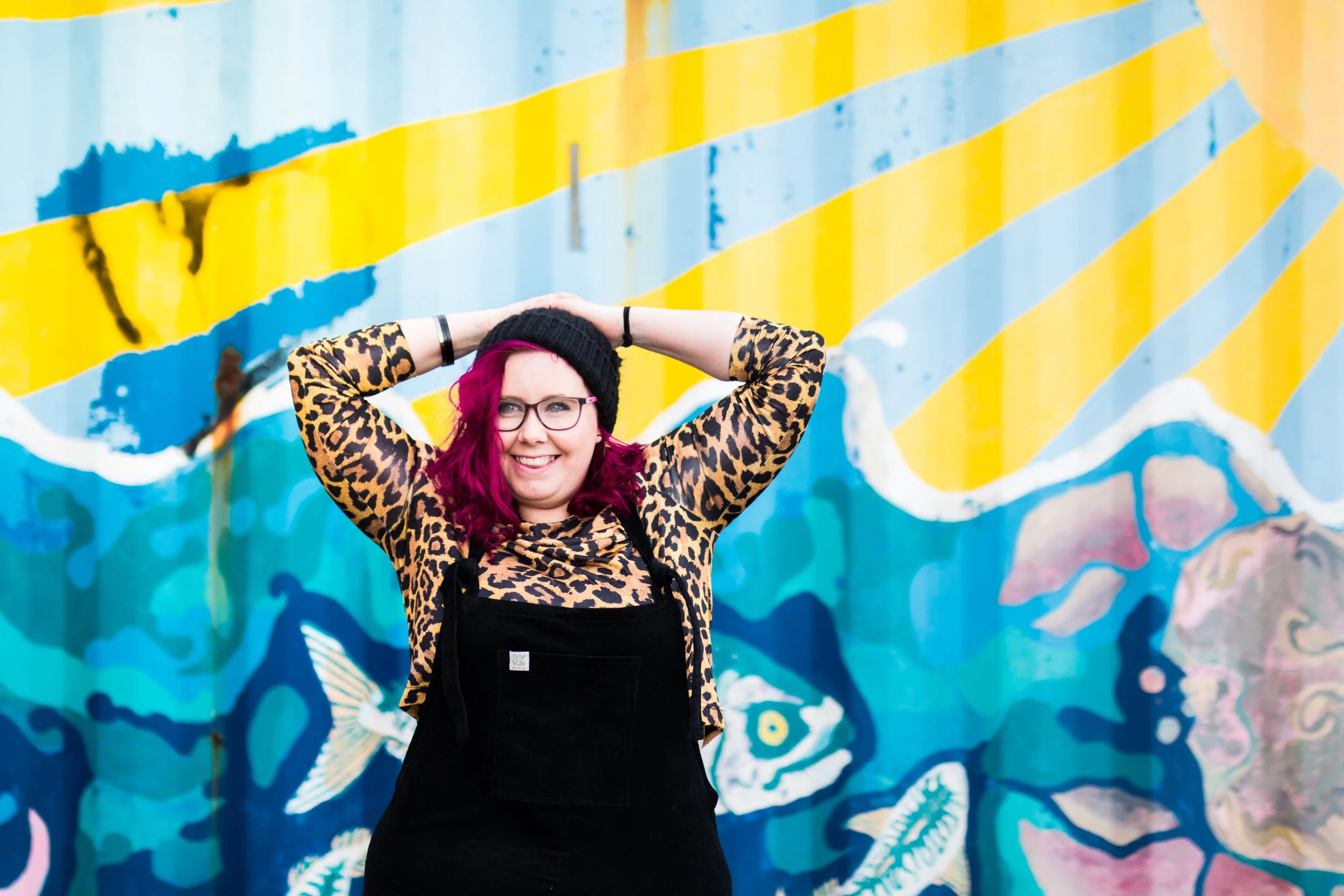 Cariad Martin, a smiling woman with pink hair, wearing glasses, a leopard print top, a black beanie, and black overalls, standing in front of a colorful mural with blue, yellow, and pink hues.