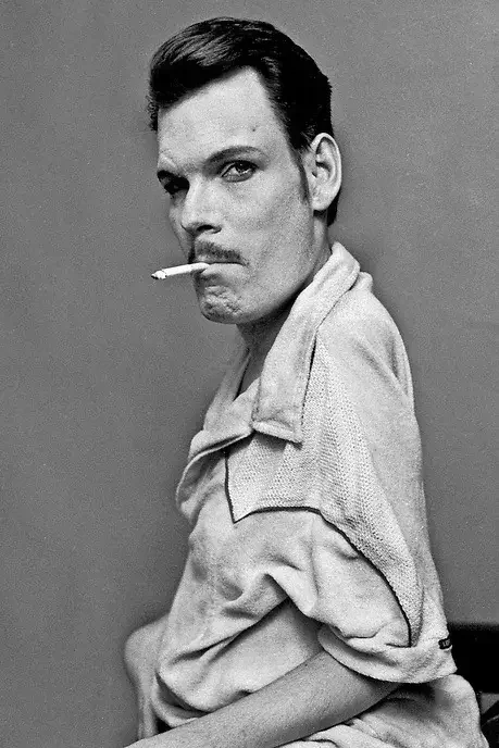 A black-and-white photo of a man with slicked-back hair, smoking a cigarette, wearing a light-colored collared shirt, and looking at the camera with a serious expression.