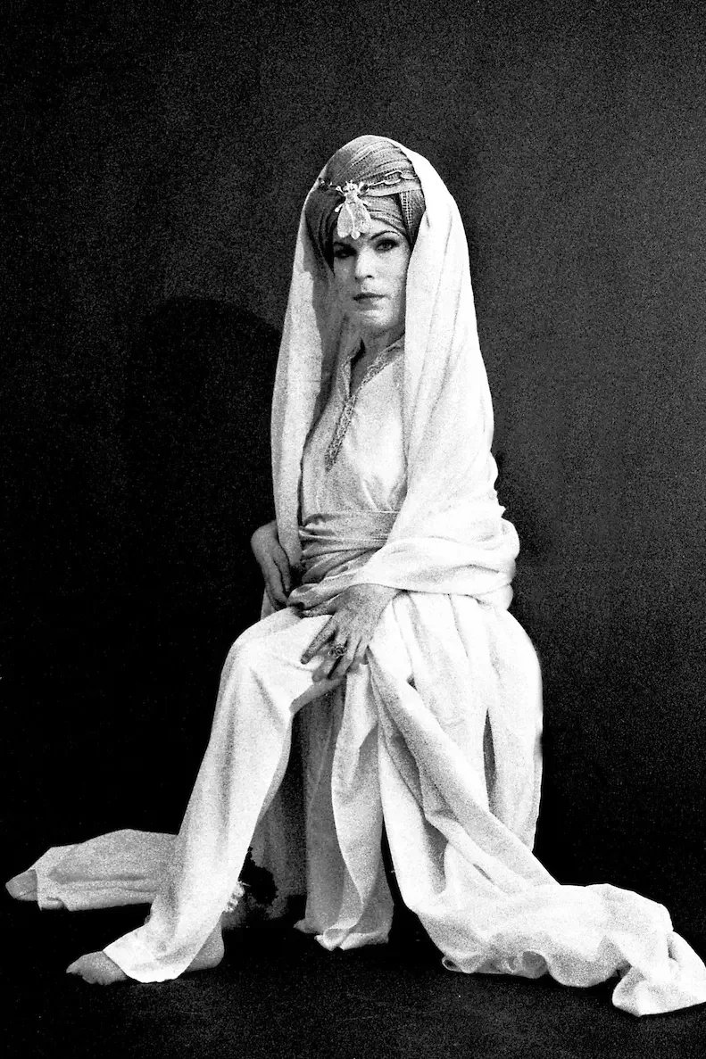 A woman dressed in elegant vintage clothing, sitting on a stool with a dark background, wearing a headscarf with ornate decoration, looking directly at the camera.