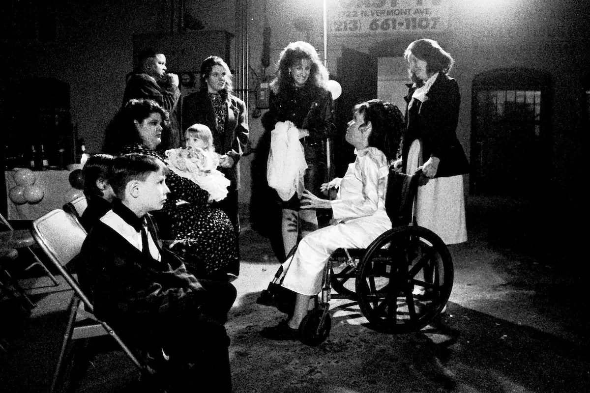 A black and white photo of a group of women and children gathered outdoors at night. A woman in a wheelchair, dressed in white, is interacting with another woman seated nearby. Several children are sitting or standing around, and one woman is holding
