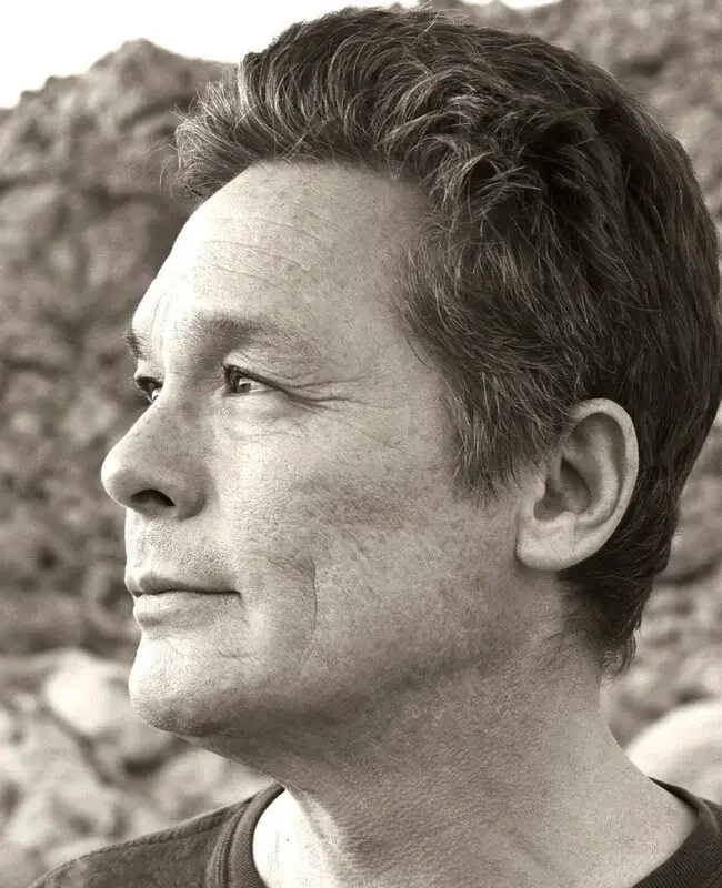 Profile of a middle-aged man with short hair, looking to the left, outdoors with rocky background, in black and white.