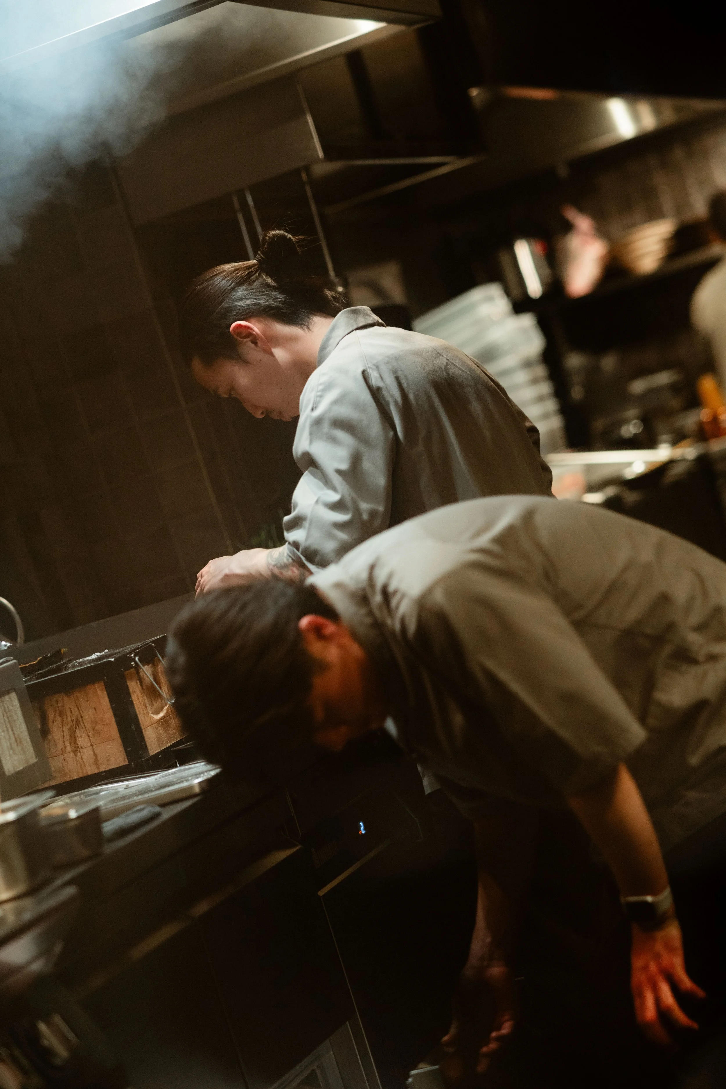Two chefs leaning over a counter in a Japanese Izakaya-style kitchen, with dark cabinets, shelves, and various kitchen items in the background, under warm lighting.