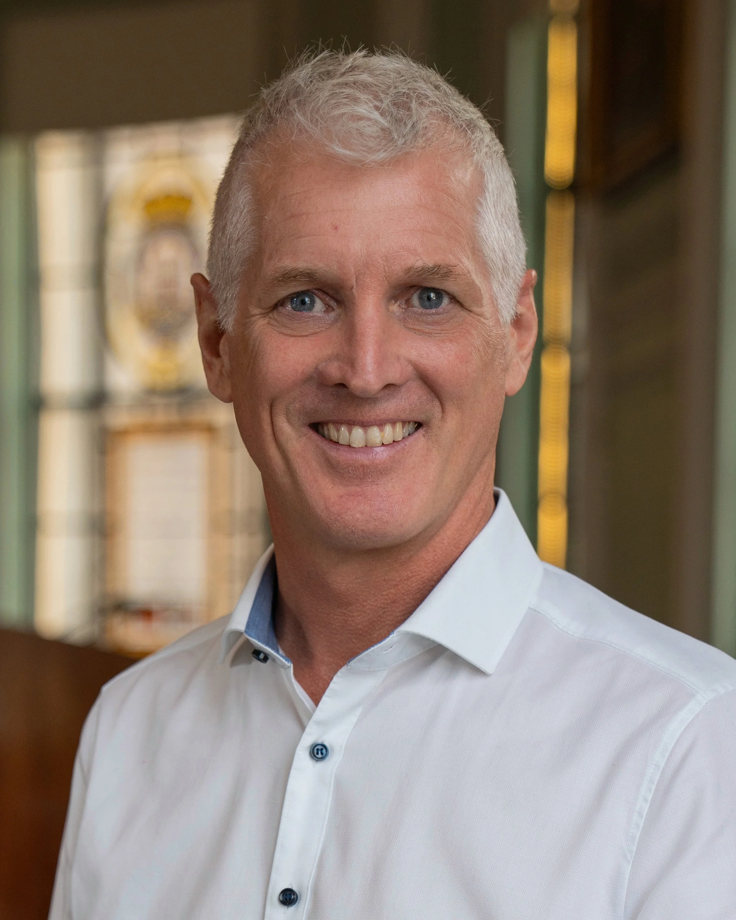 Close-up of a smiling man who is Neil Pendle with short, grey hair, wearing a white dress shirt with a light blue collar, in an indoor setting.