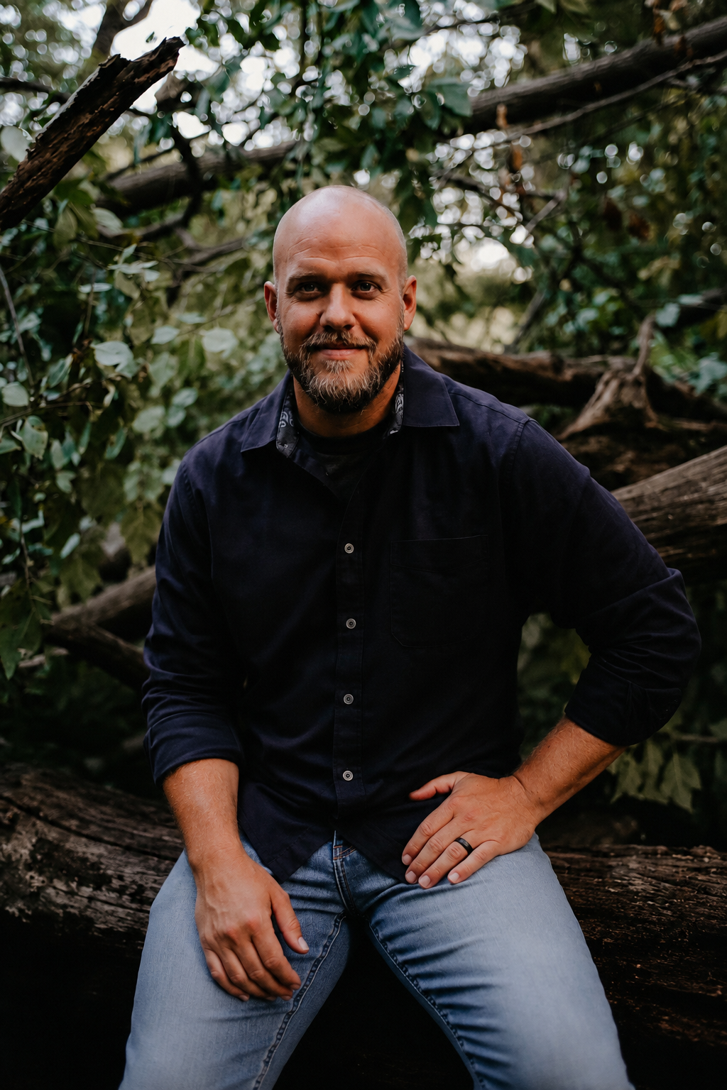A man with a shaved head, beard, and mustache is sitting on a fallen tree trunk outdoors in a wooded area with green leaves and branches in the background. He is wearing a dark navy shirt and light blue jeans, with his right hand resting on his thigh and his left hand on his hip.