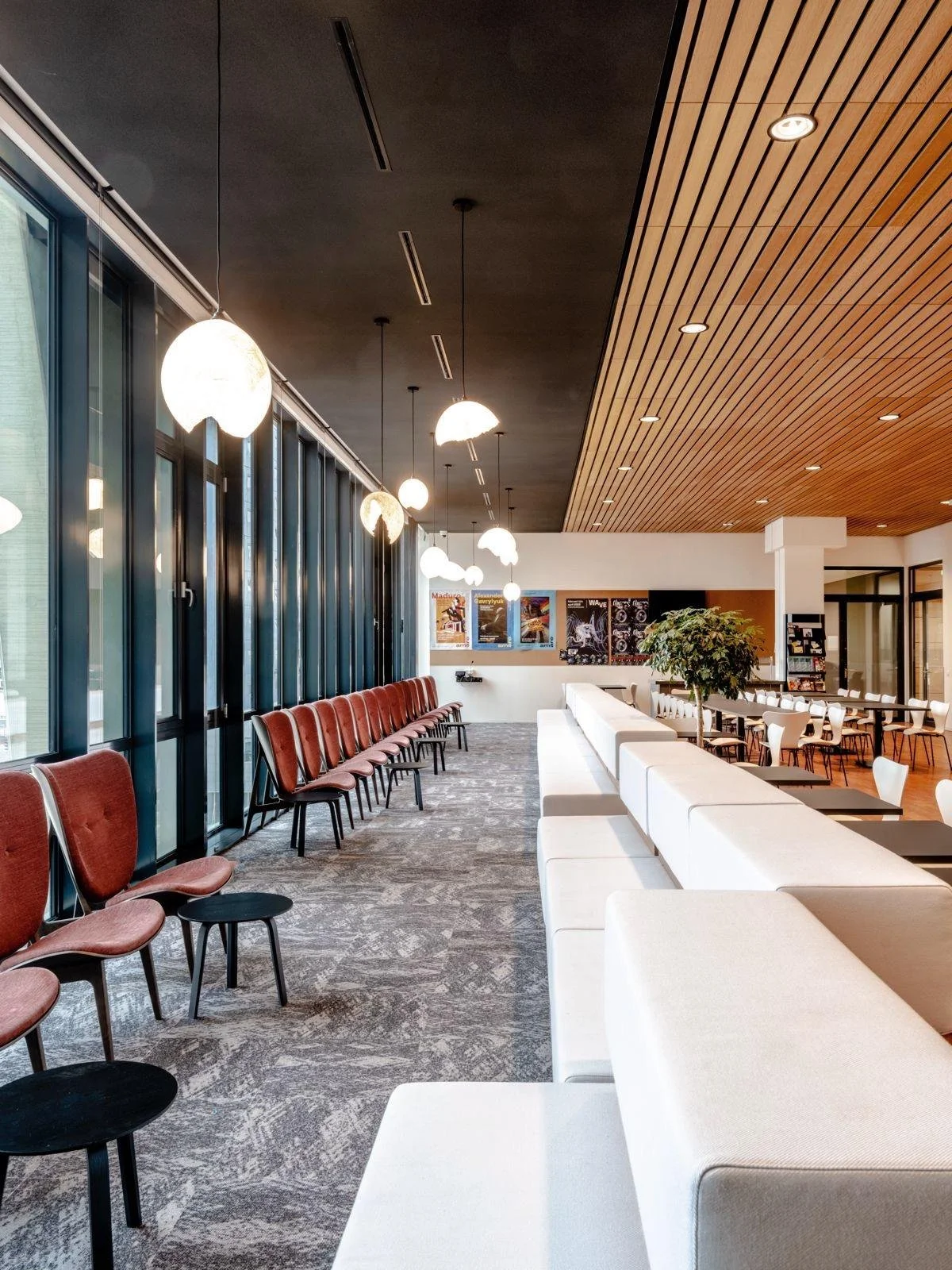 Interieur van een modern gemeenschapsruimte of lounge met rijen rode en witte stoelen, grote ramen met gordijnen, hangende plafondlampen en posters aan de muur, inclusief een boom in een plantenbak.