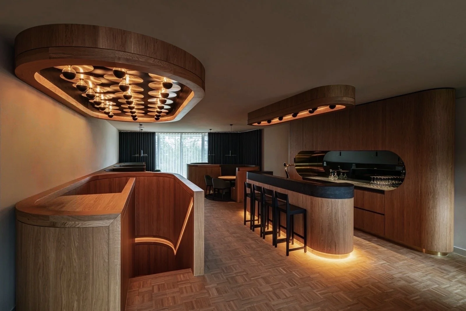 Interieur van een modern café of bar met houten inrichting, elegante verlichting, bar met krukken en een zitgedeelte met tafel en stoelen, grote ramen met gordijnen.