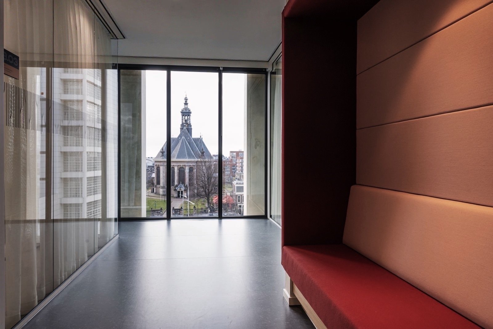 Binnenzicht van een modern kantoor met grote ramen die uitzicht bieden op een historische kerk en moderne gebouwen, een zithoek met een gebogen rood bankje aan de rechterzijde.