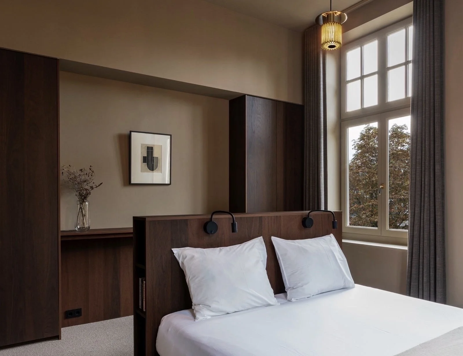 Een slaapkamer met een bed, wit beddengoed en twee witte kussens, grote ramen met grijze gordijnen, een wandlamp boven het hoofd van het bed, een kunstwerk aan de muur en een vaas met gedroogde bloemen op een plank.
