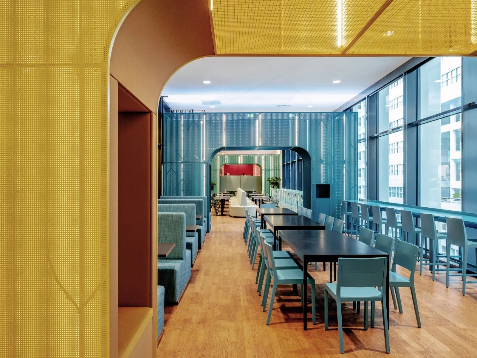 Een moderne, kleurrijke eetzaal met grote ramen, blauwe en gele wanden, en verschillende zitplaatsen, waaronder tafels met stoelen en een aangrenzend loungegedeelte.