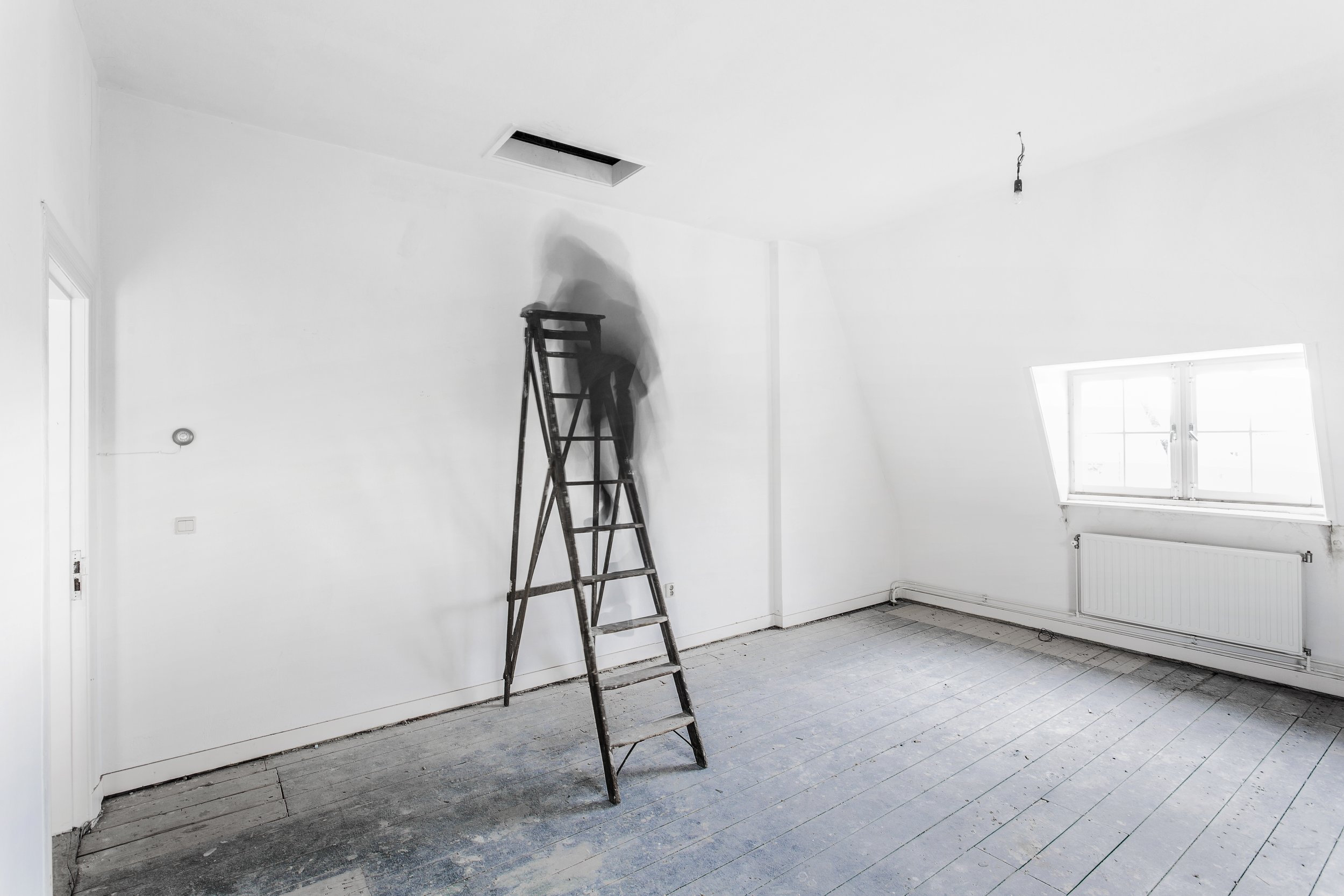 Een lege witte kamer met een ladder en een persoon die op de ladder staat, die mogelijk werkzaamheden verricht aan het plafond. Er is een raam en een radiator zichtbaar.