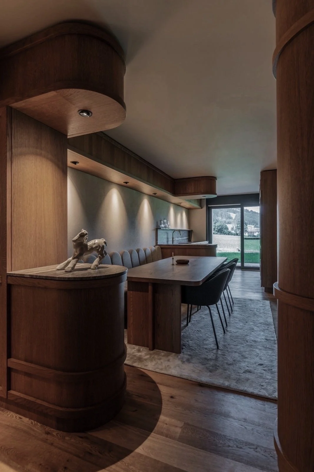 Interieur van een woonkamer met houten elementen, een grote houten eettafel, zwarte stoelen, decoratieve items, zachte verlichting, een tapijt en een groot raam dat uitzicht biedt op een groene buitenruimte.