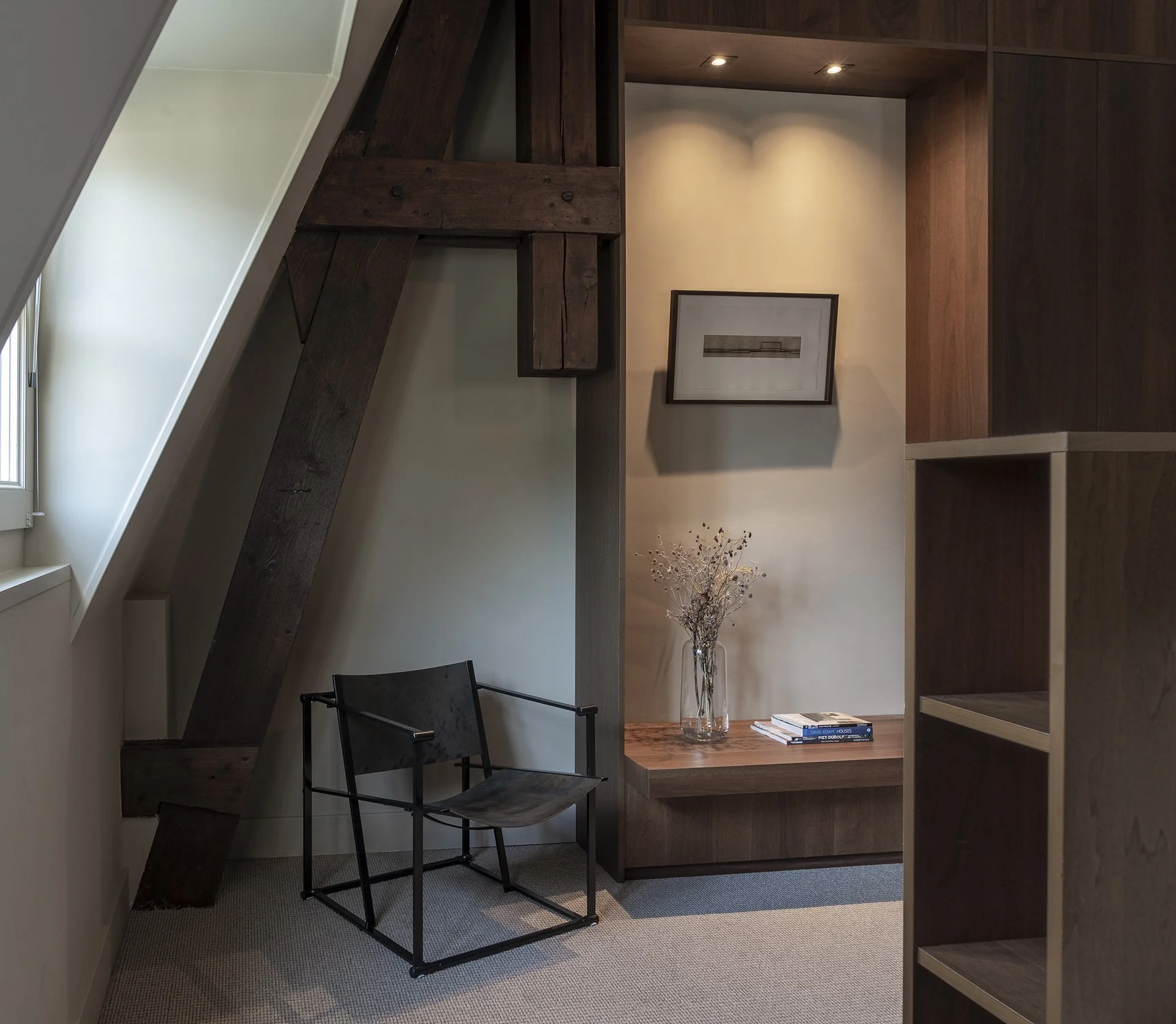 Een studeerhoek in een huis met boeken, een zwart metalen stoel, een vaas met takken en een houten muur met ingebouwde verlichting.