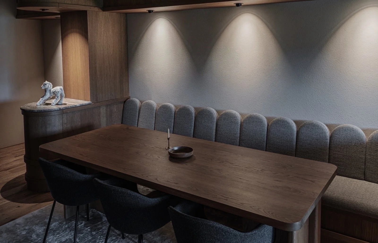 Eethoek met een grote houten tafel, vier zwarte stoelen, en een beige bank met een gebogen achterkant. Aan de muur hangen drie inbouwlampen die lichte op de muur werpen. Op de tafel ligt een klein vaasje met een kaars en een houten bord. Aan de linke