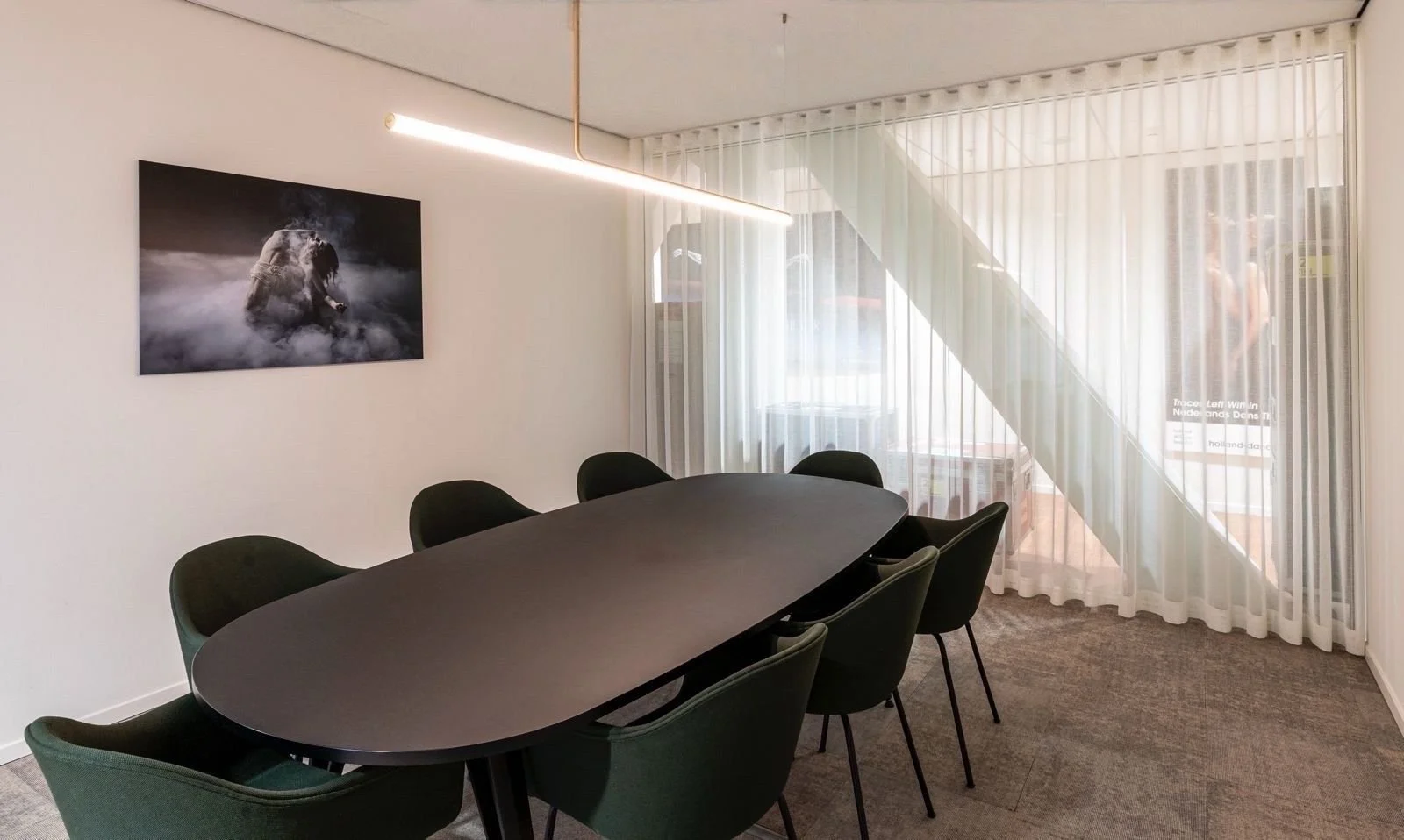 Een vergaderruimte met een zwart ovalen tafel en acht zwarte stoelen. Aan de muur hangt een zwart-wit foto van een persoon die in rook omgeven is. Grote ramen met witte vitrage laten daglicht binnen.