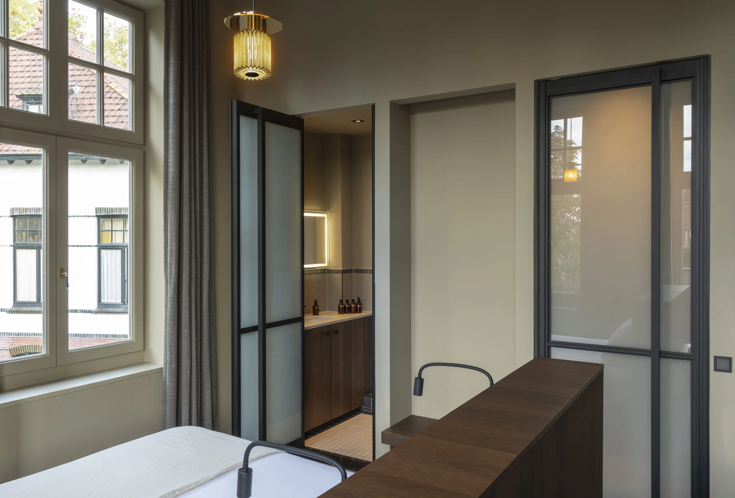 Een kamer met grote ramen, een bed met wit beddengoed, een houten bureau, een scheidingswand met zwarte metalen kader, een badkamer met een spiegel en een ladenkast, moderne verlichting.
