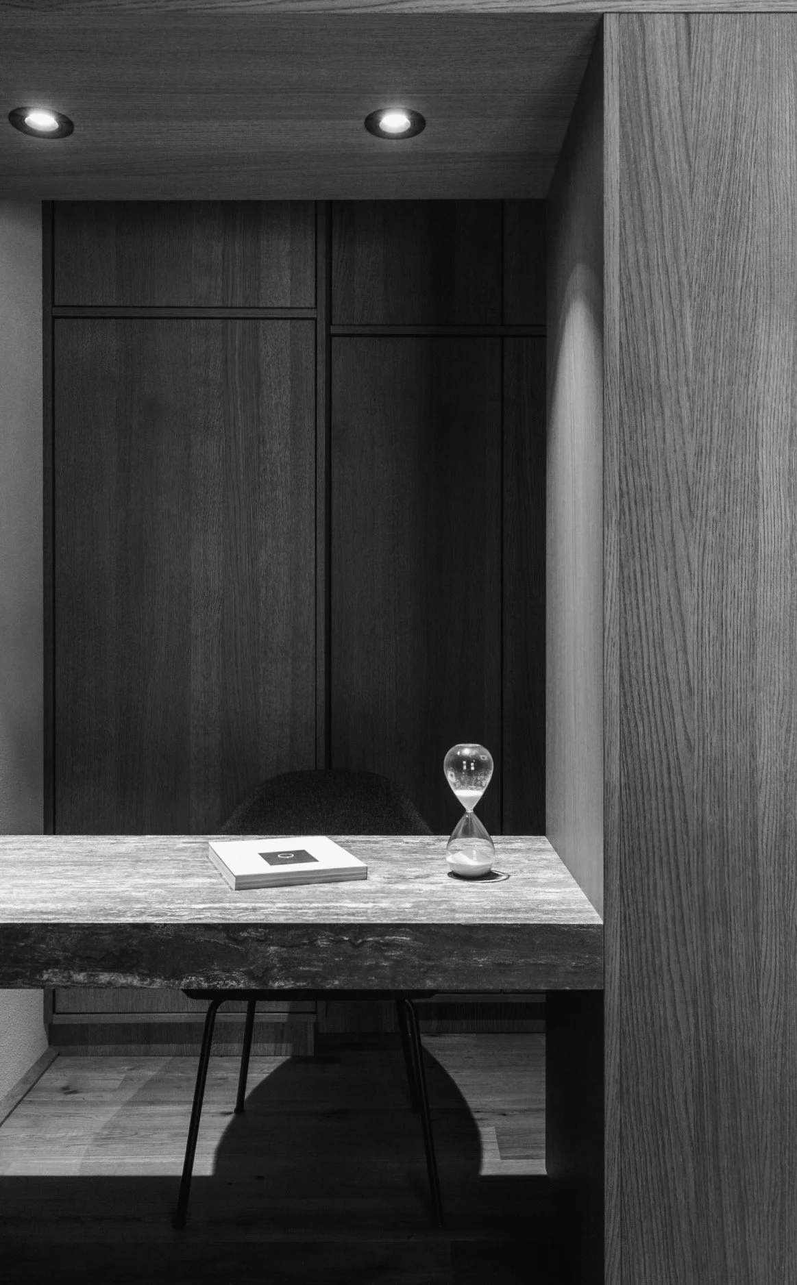 Een zwart-witten afbeelding van een interieur met een houten bureau, een boek, een zandloper en een stoel. Achter het bureau bevinden zich houten kasten met een plafondverlichting.