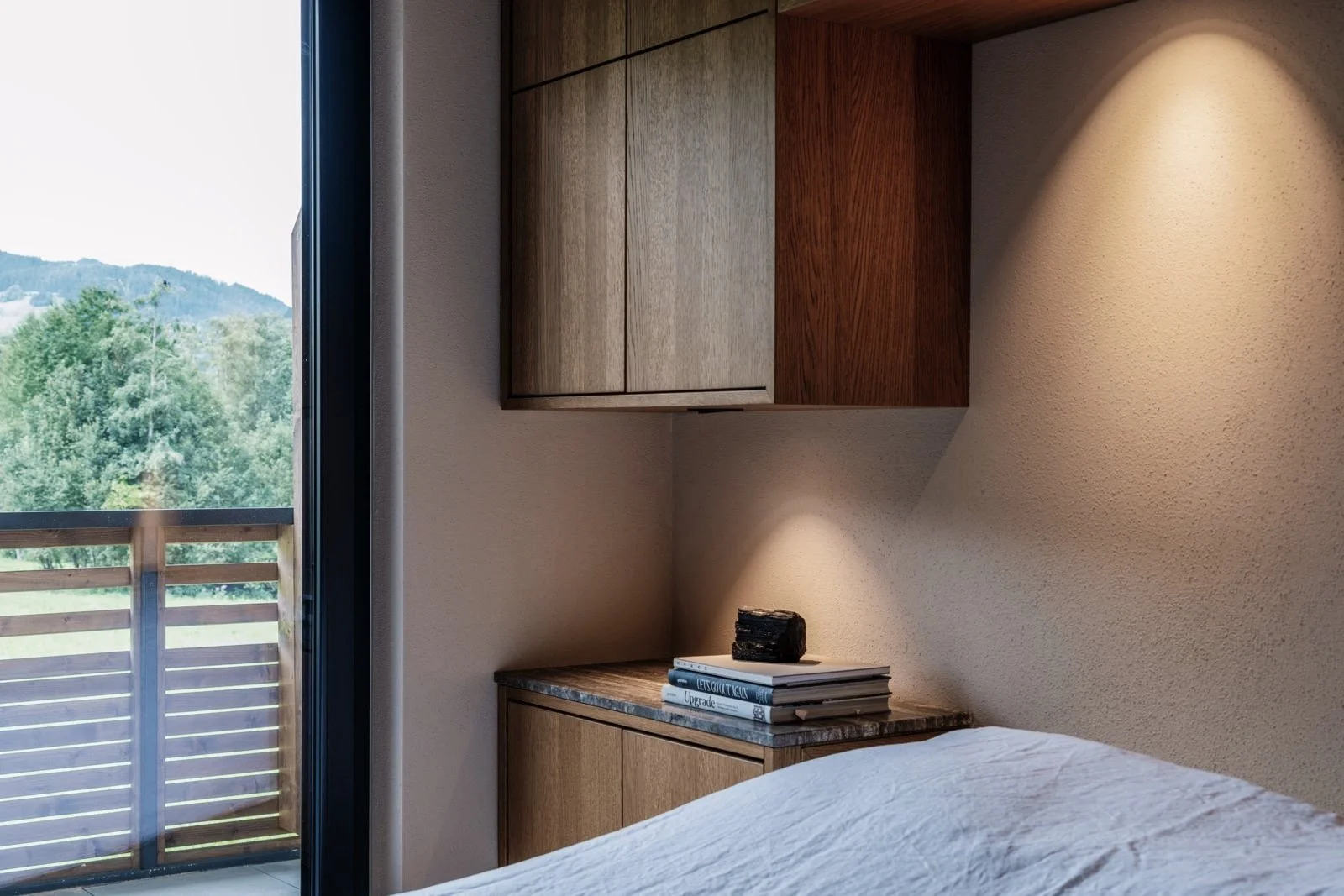 Een slaapkamer met een bed en een nachtkastje naast een groot raam dat uitkijkt op een bos en bergen op de achtergrond.