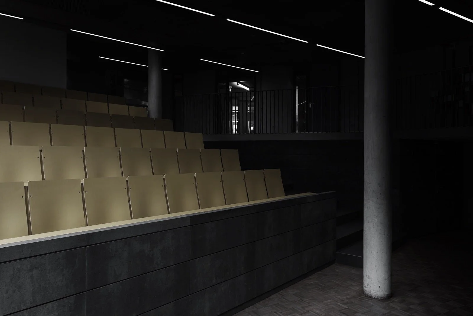 Een zaal met lege beige stoelen en donkere muren, enkele lichtstrips aan het plafond, en een betonnen pilaar voor een modern, minimalistisch interieur.