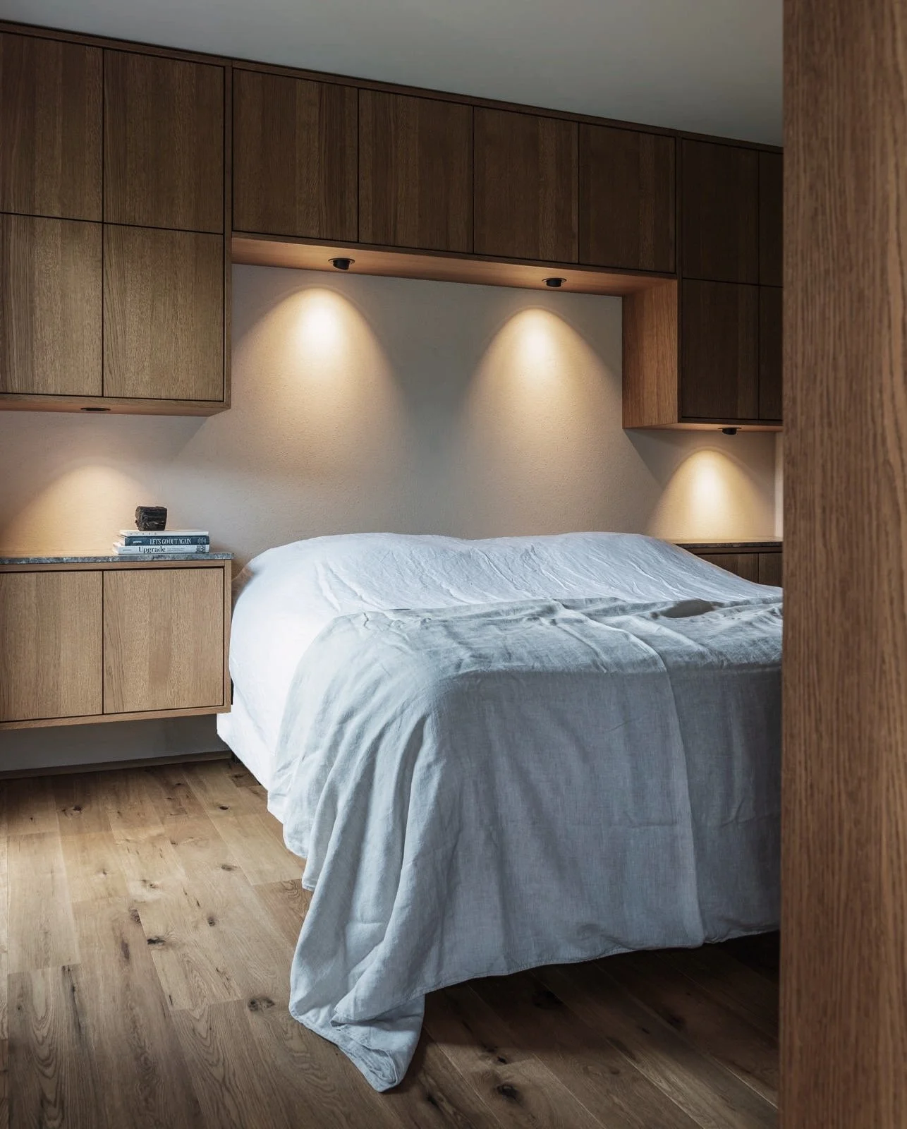Een slaapkamer met een bed dat bedekt is met een lichtgrijze bedsprei, houten wandkasten en ingebouwde plafondverlichting die gericht is op de muur boven het bed.