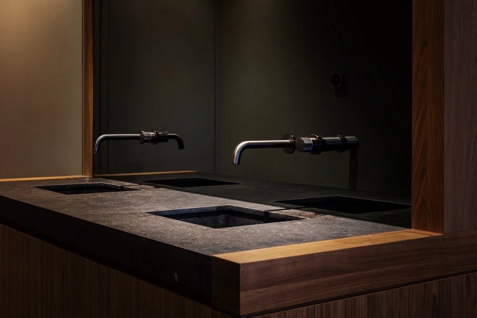 Een donkere toiletruimte met twee basins zonder kranen, een grote spiegel en houten panelen en werkblad.